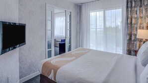 A hotel room with a large bed, a flat-screen TV mounted on the wall, a sliding glass door leading to a balcony, and a view of the outdoors. - Quality Suites (Ste Anne de Beaupre)