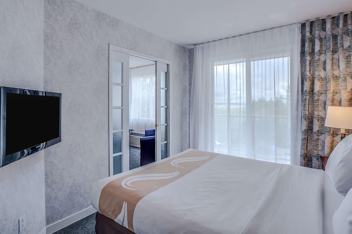 A hotel room with a large bed, a flat-screen TV mounted on the wall, a sliding glass door leading to a balcony, and a view of the outdoors.
