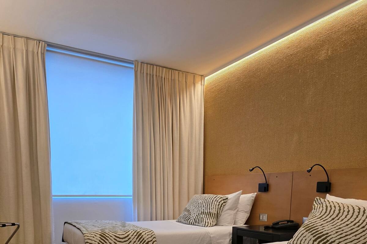 Habitación de hotel con dos camas, un ventanal con cortinas, cabecero con iluminación integrada y suelo de madera.