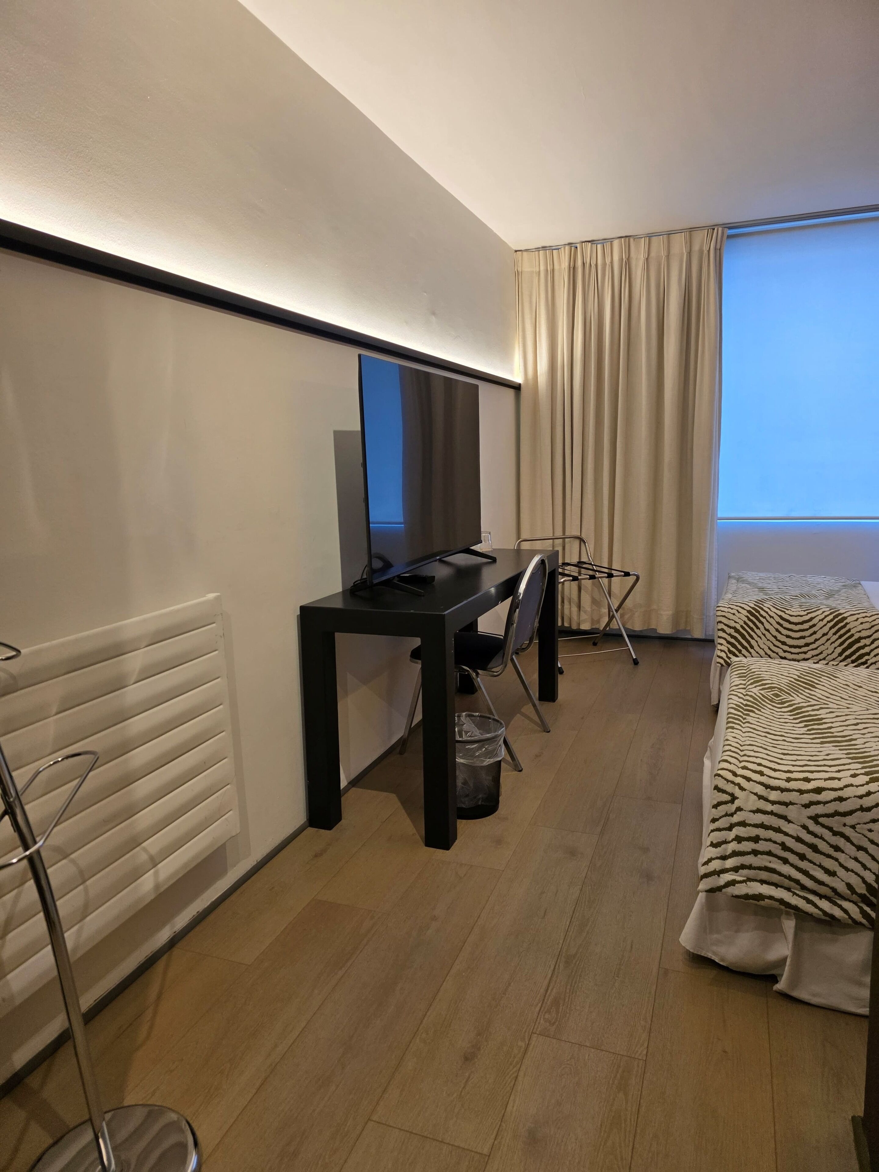 A hotel room with a bed, a desk with a computer, a chair, a radiator, and a window with curtains.