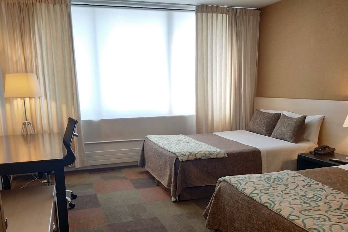 Habitación de hotel con dos camas, un escritorio y una silla. Cuenta con una ventana con cortinas, una lámpara y suelo alfombrado.