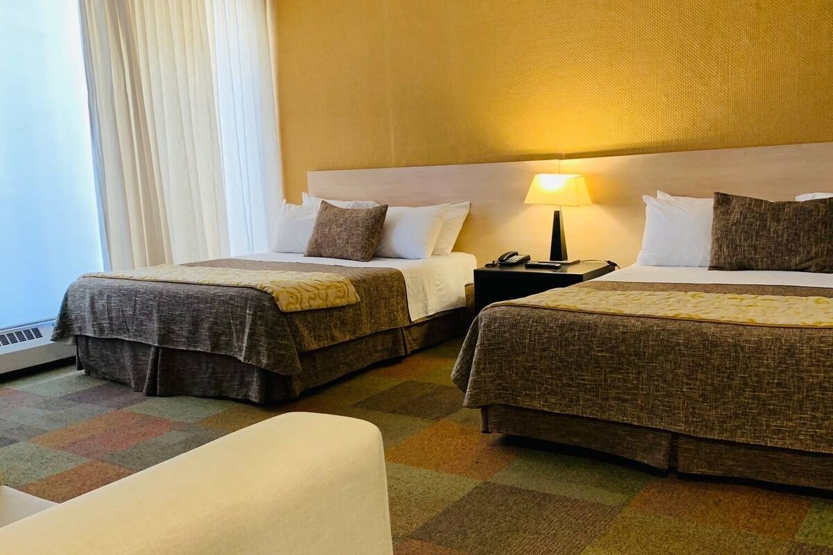 Habitación de hotel con dos camas, un sofá, una mesita de noche con lámpara y un ventanal con cortinas.
