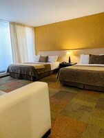 Habitación de hotel con dos camas, un sofá, una mesita de noche con lámpara y un ventanal con cortinas.