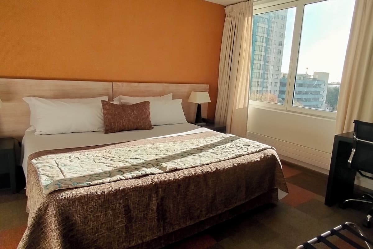 Habitación de hotel con una cama grande, dos mesitas de noche con lámparas, una silla y vistas a la ciudad desde la ventana.