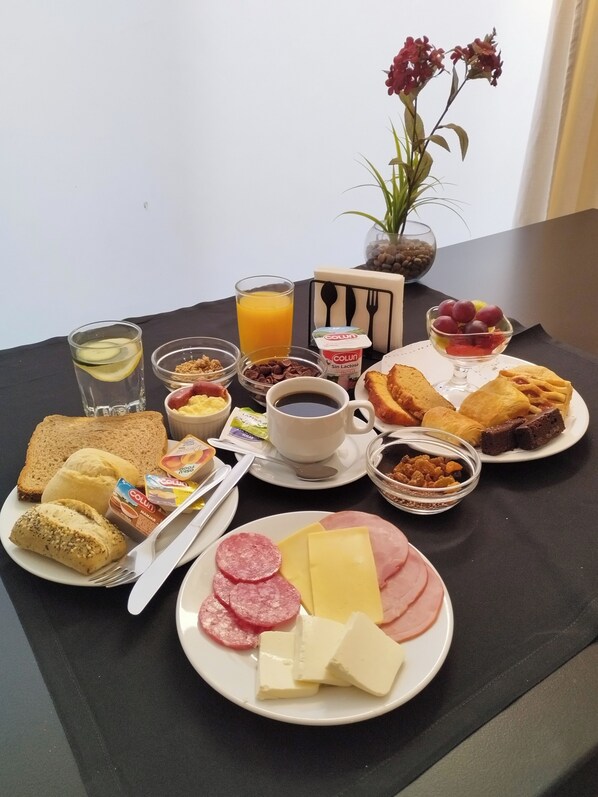 A breakfast table with various food items including toast, bread rolls, cheese, ham, sausages, and a fruit bowl. There are also drinks, a coffee cup, and a small plant in the background.