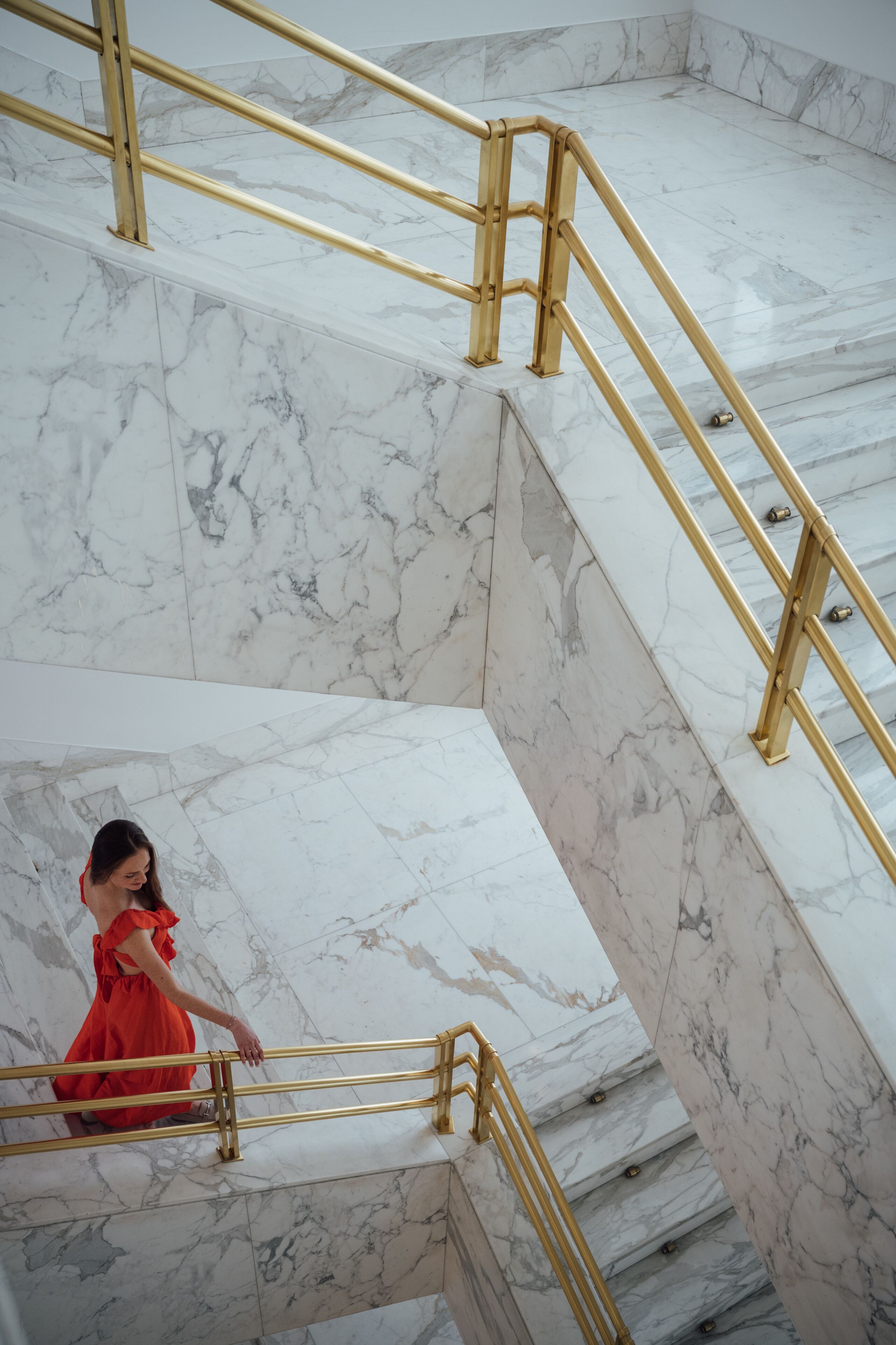 A marble staircase with golden railings and a person in a red dress descending.