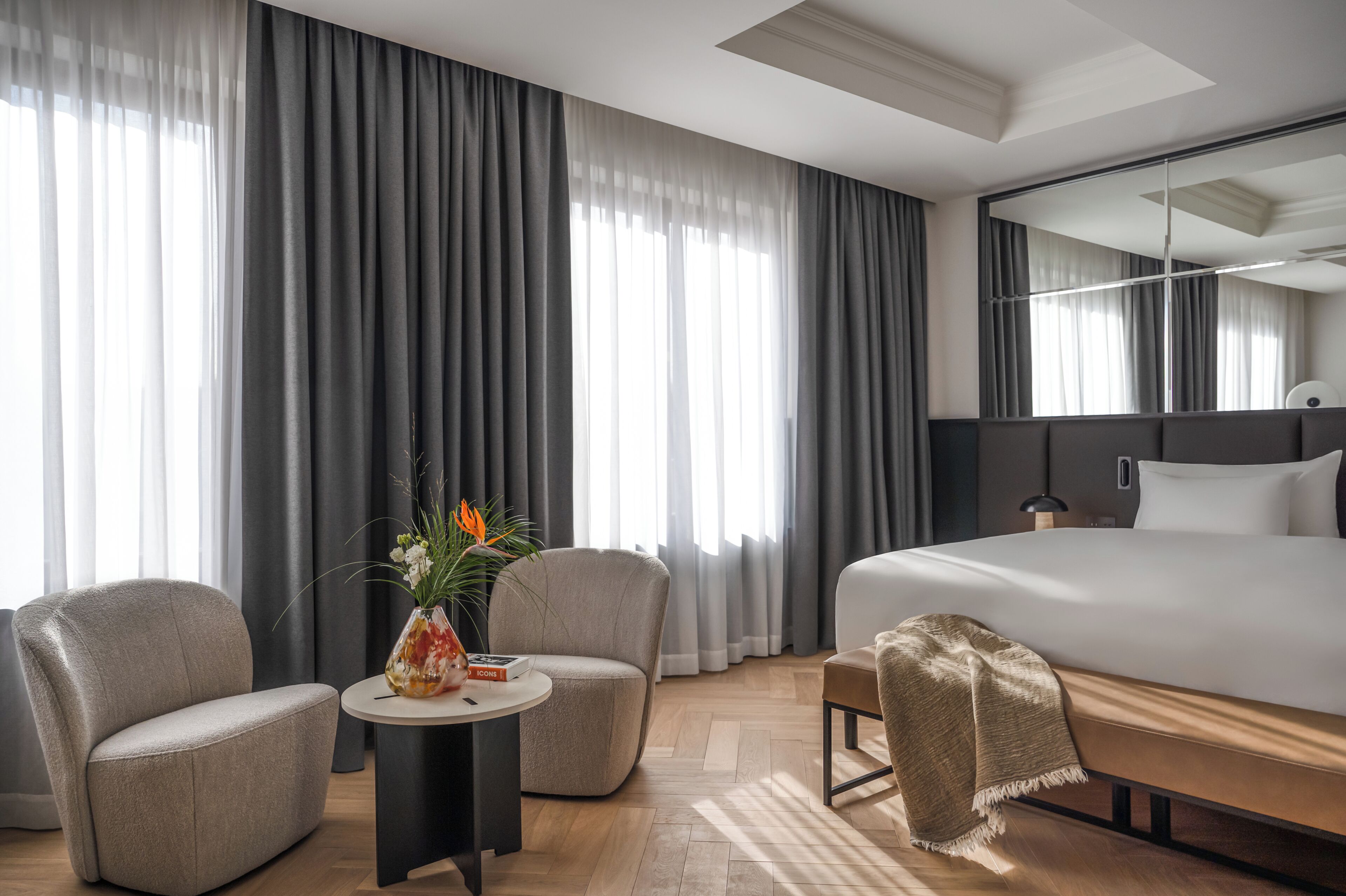 Habitación de hotel con una cama grande, dos sillones, una mesita con un florero y un ventanal con cortinas.