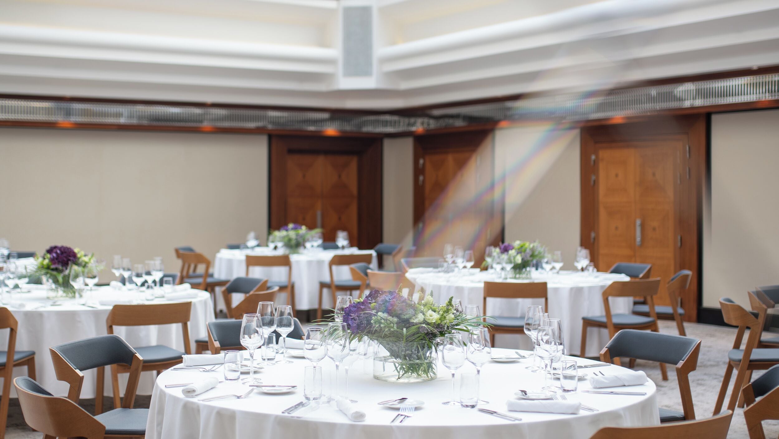 A banquet hall with round tables set for dining, each with a white tablecloth, chairs, and glassware.