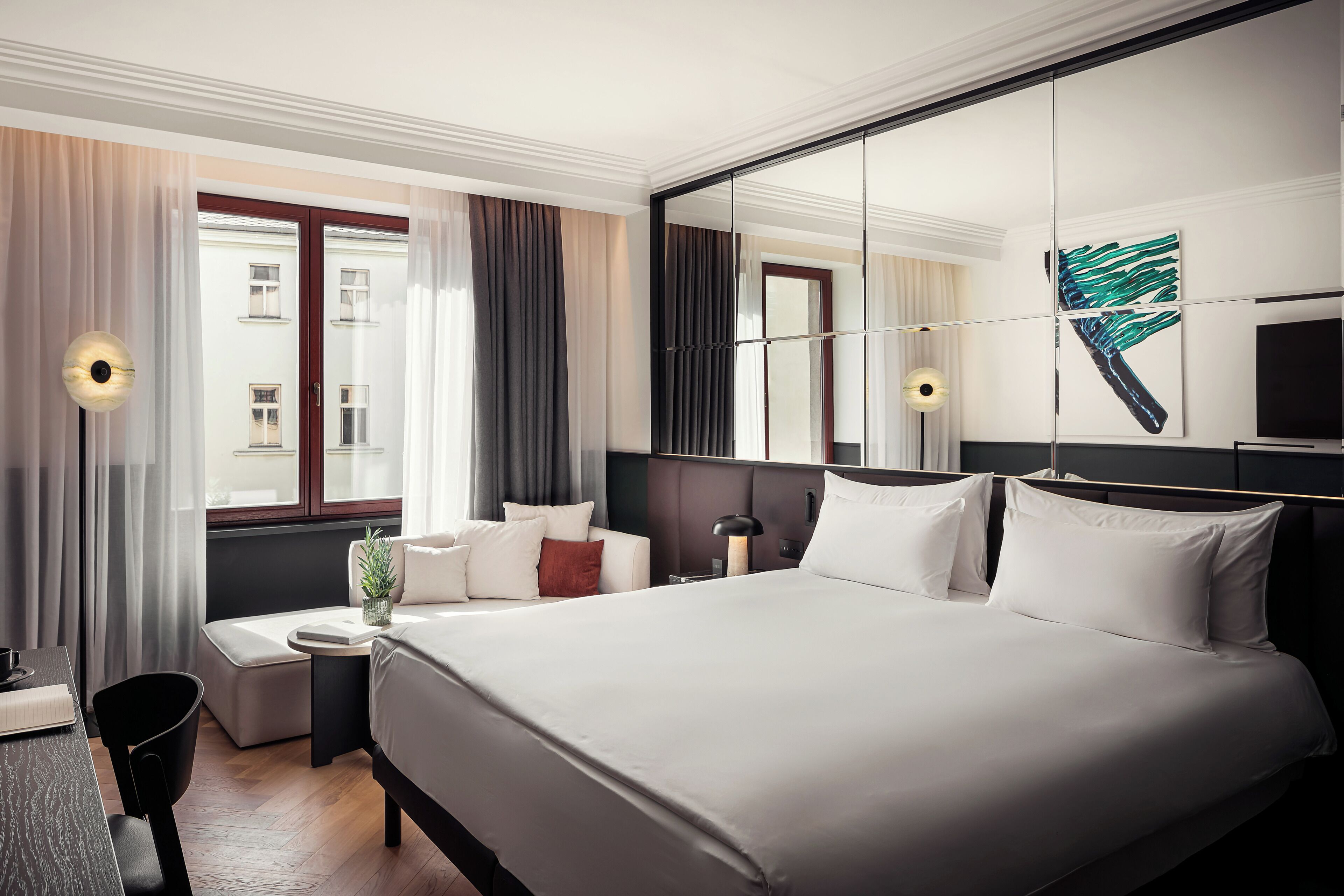 Una habitación de hotel moderna con una cama grande, una mesita de noche, una silla, una pequeña mesa, una planta en maceta y un espejo amplio.