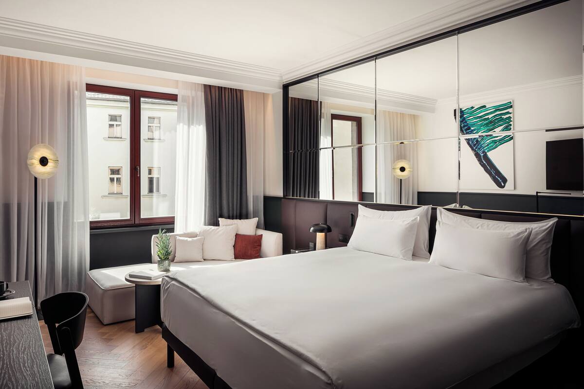Una habitación de hotel moderna con una cama grande, una mesita de noche, una silla, una pequeña mesa, una planta en maceta y un espejo amplio.