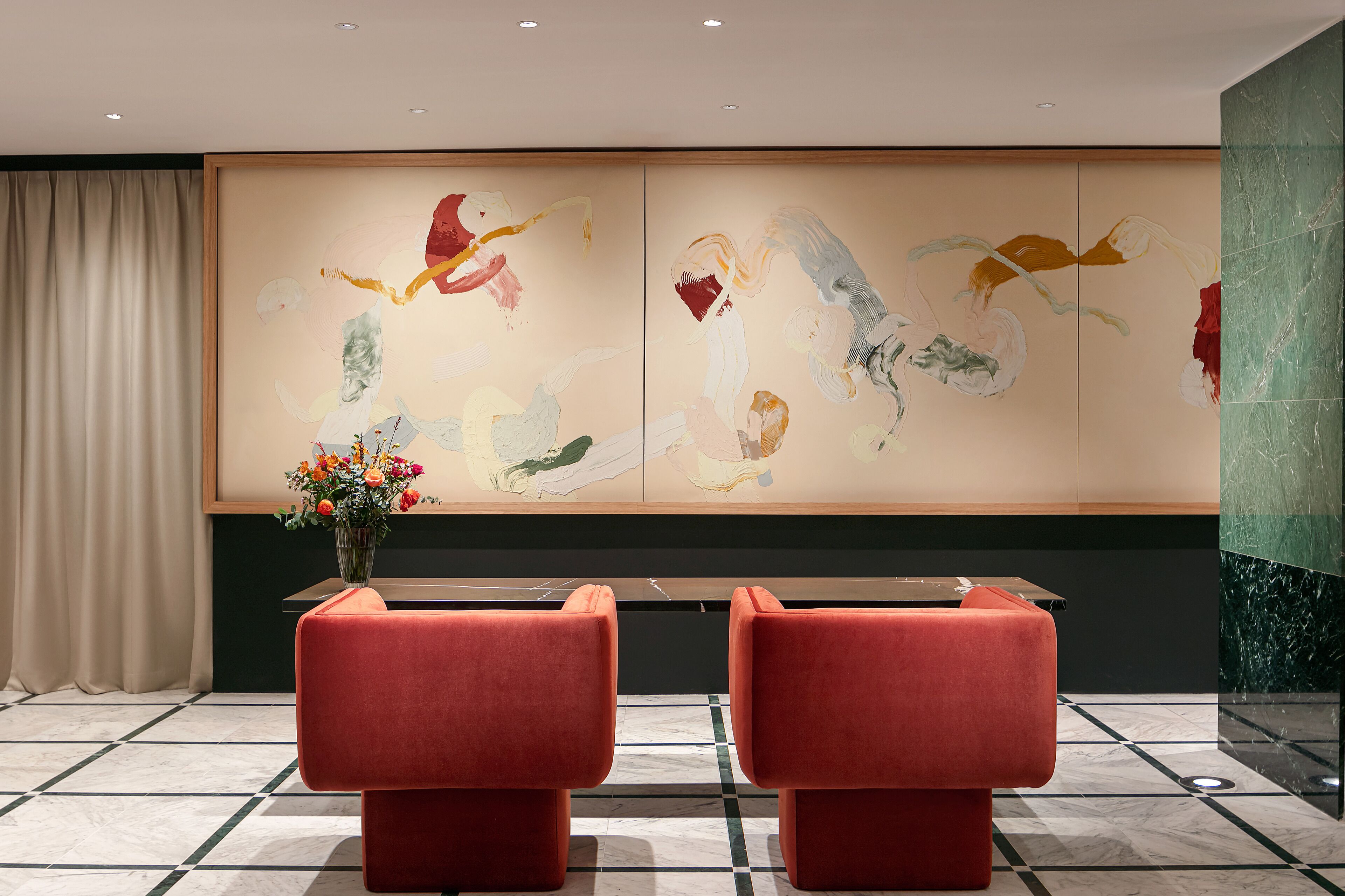 A lobby with a large abstract painting, two red chairs, a vase with flowers, and a marble floor.