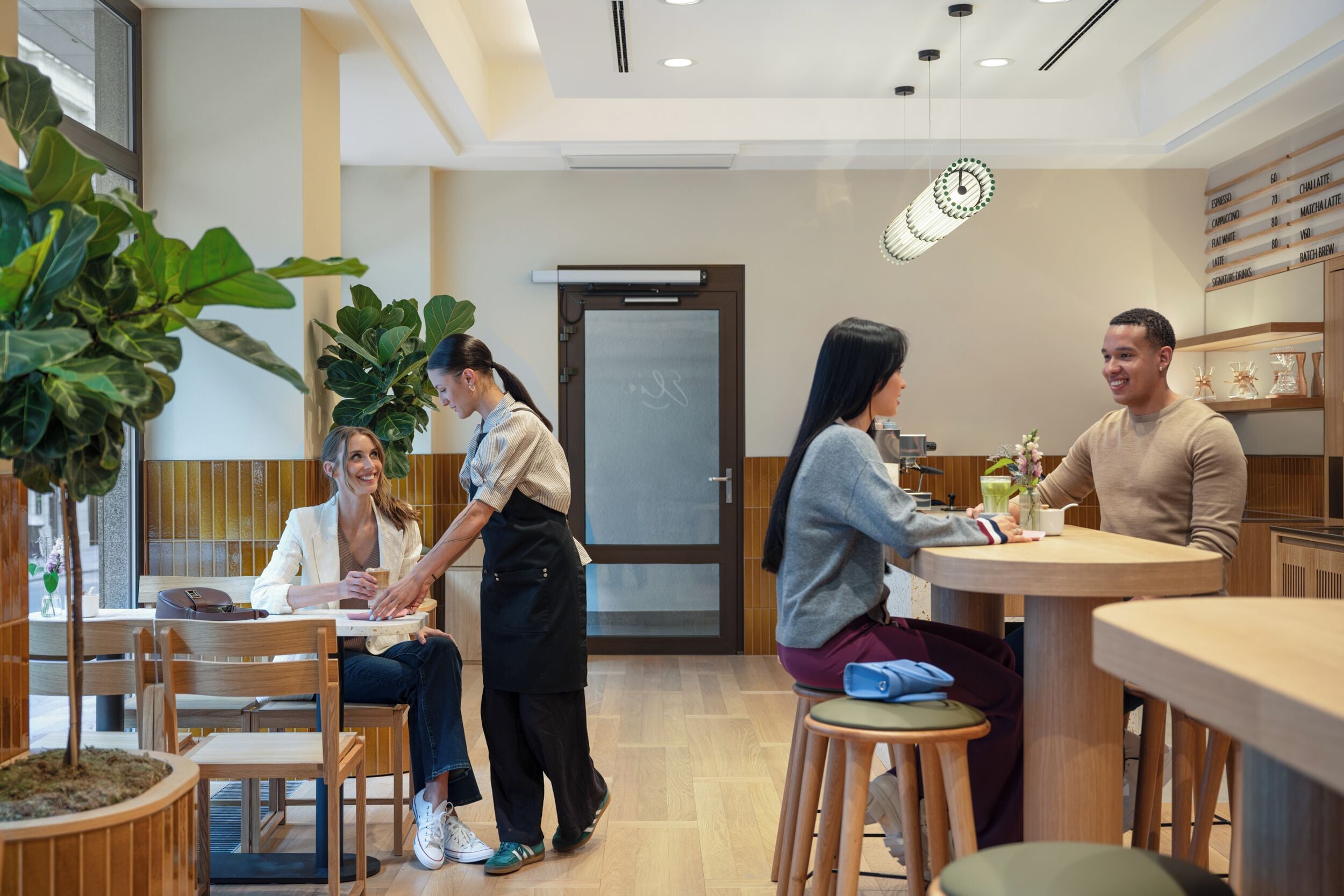 A modern cafe with wooden furniture, large potted plants, and a menu board on the wall.