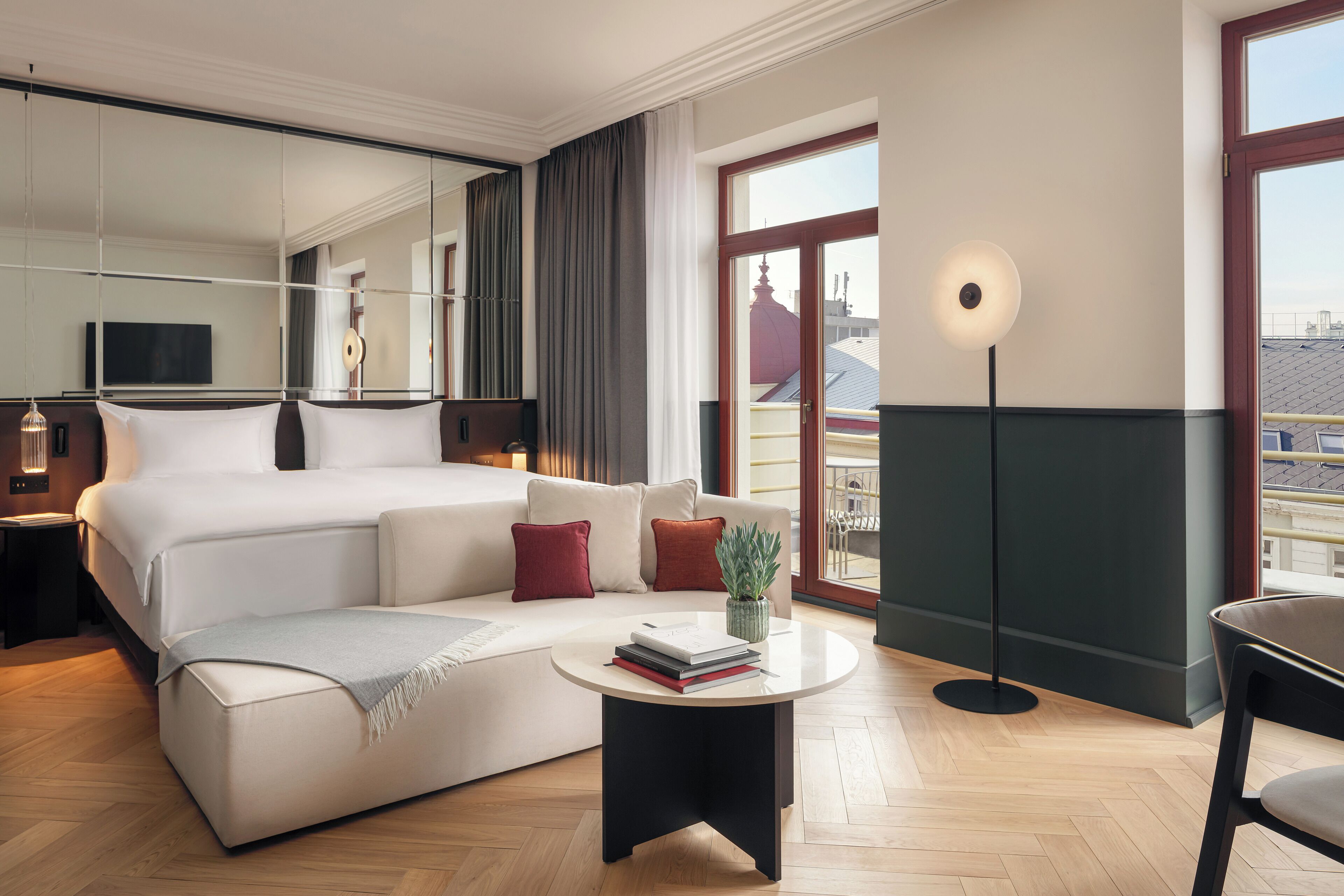 Habitación de hotel moderna con una cama grande, un sofá, una mesa de centro redonda y vistas de la ciudad a través de amplios ventanales.