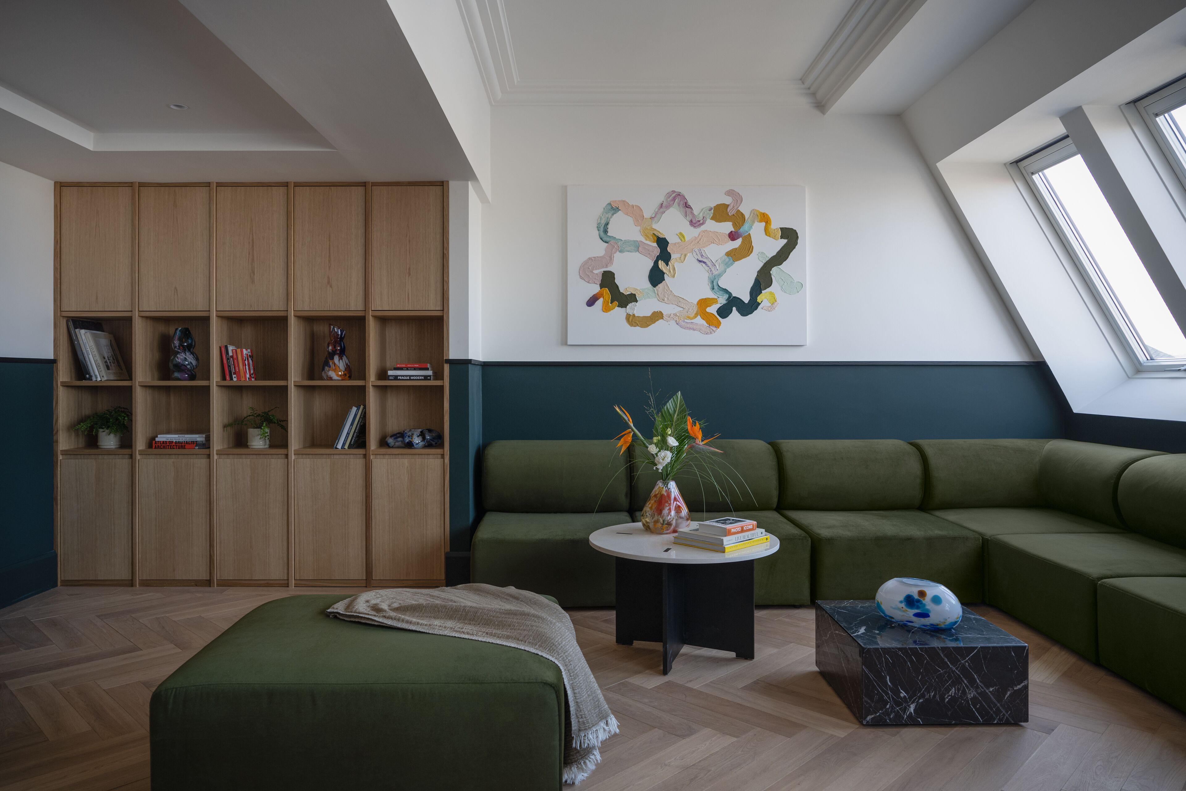 A modern living room with a green sofa, a wooden bookshelf, and a painting on the wall.