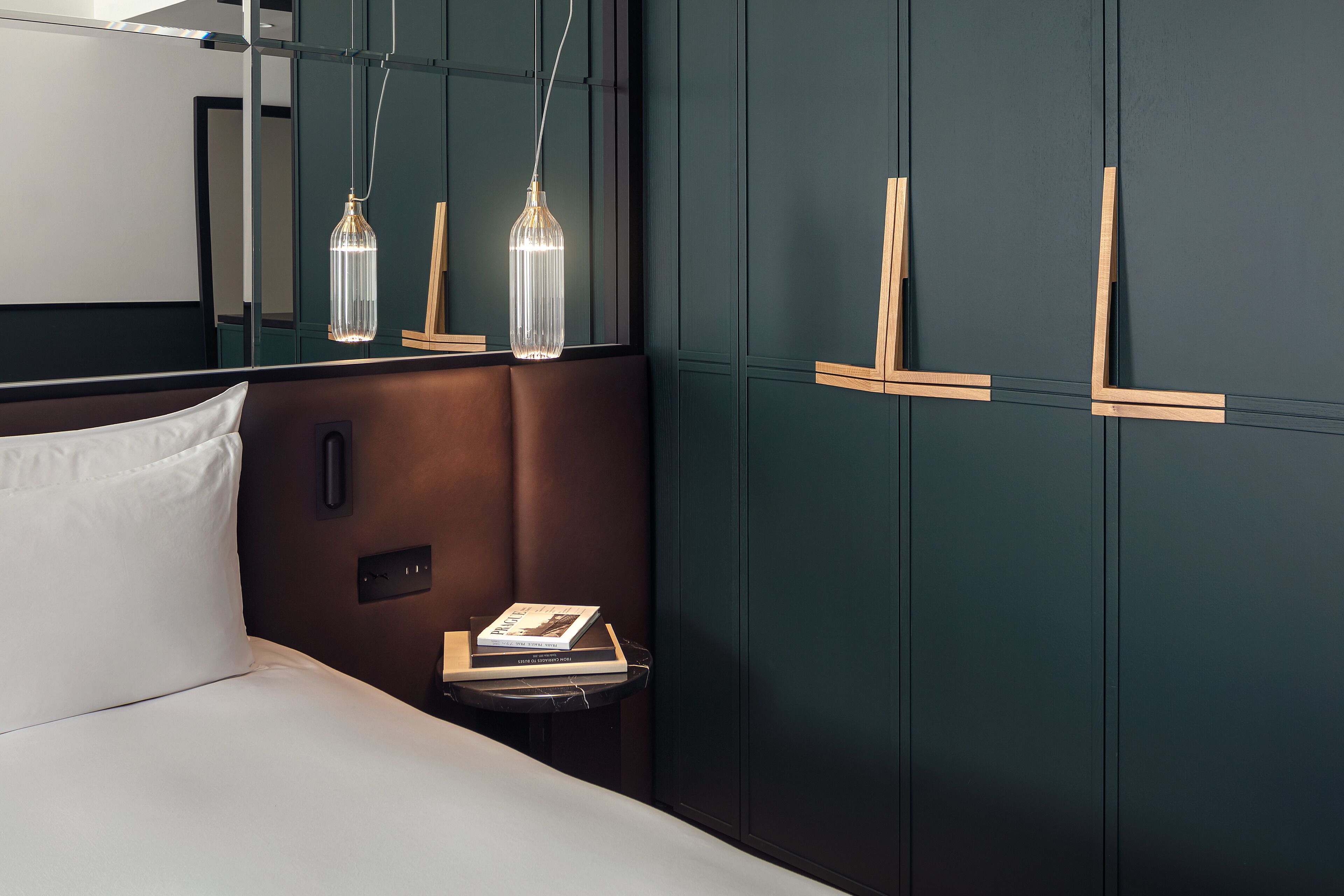 Ein modernes Hotelzimmer mit einer dunkelgrünen Wand, einem hölzernen Kopfteil, einem weißen Bett und einem kleinen Nachttisch mit Büchern.