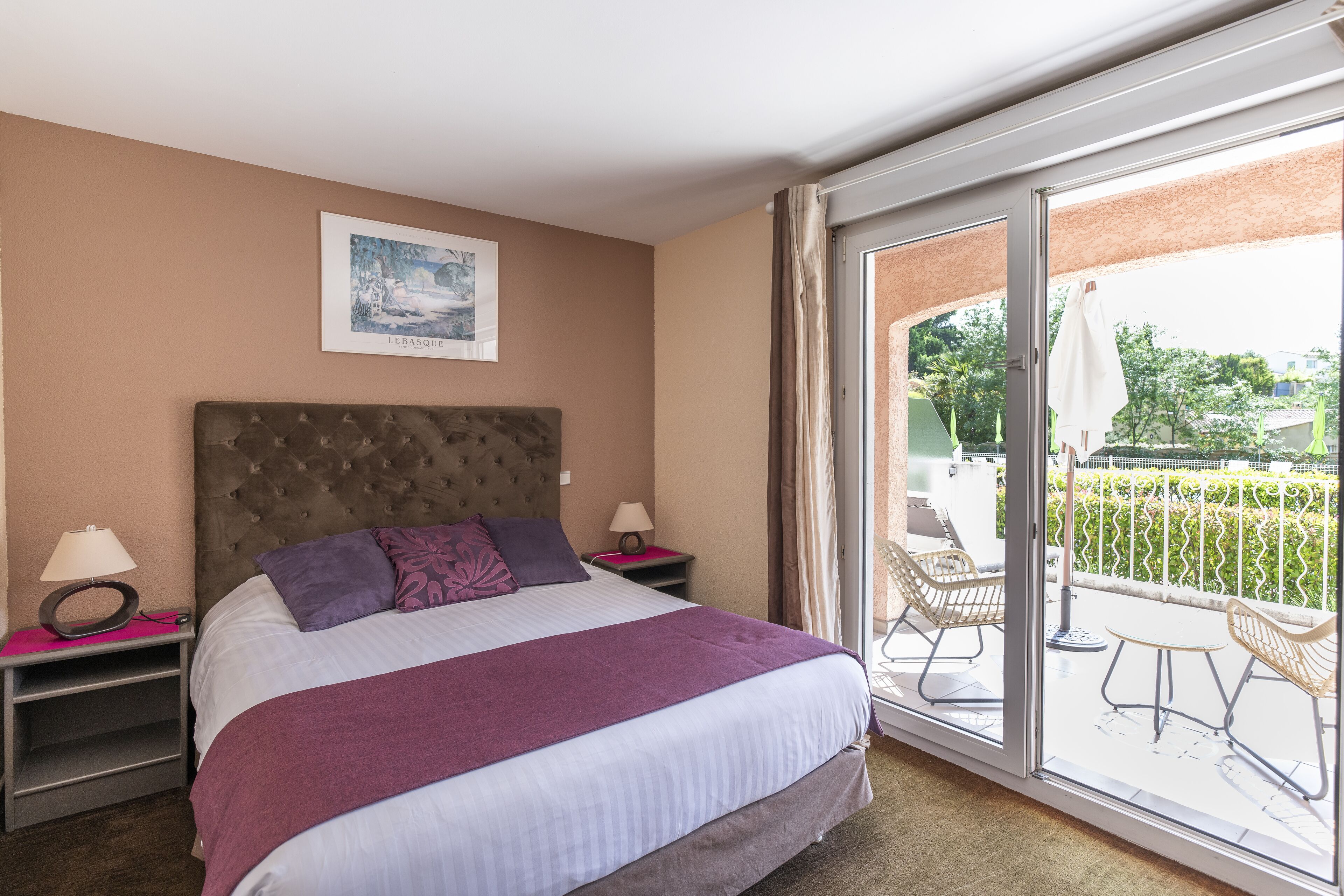 Une chambre d’hôtel avec un grand lit, une tête de lit, deux tables de chevet avec des lampes, un tableau encadré au mur et une porte-fenêtre coulissante donnant sur un espace extérieur aménagé en salon.