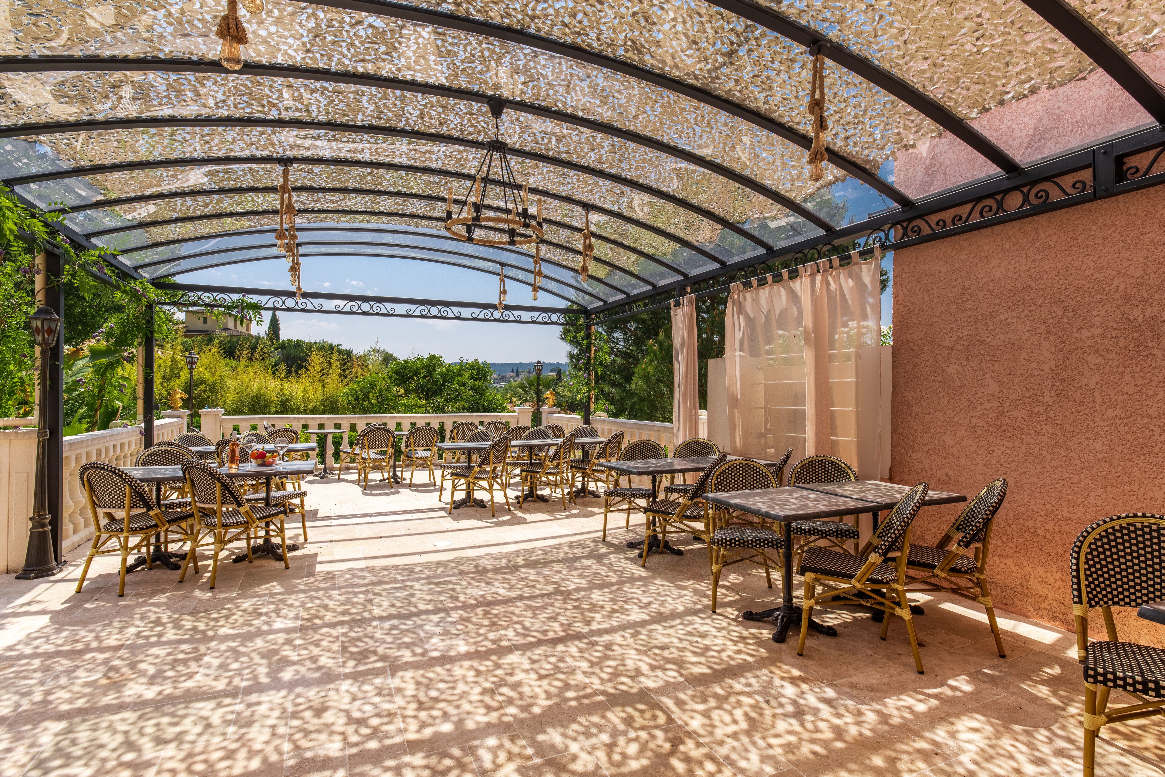 Un patio exterior con mesas y sillas, techo de vidrio y vistas a zonas verdes al fondo.