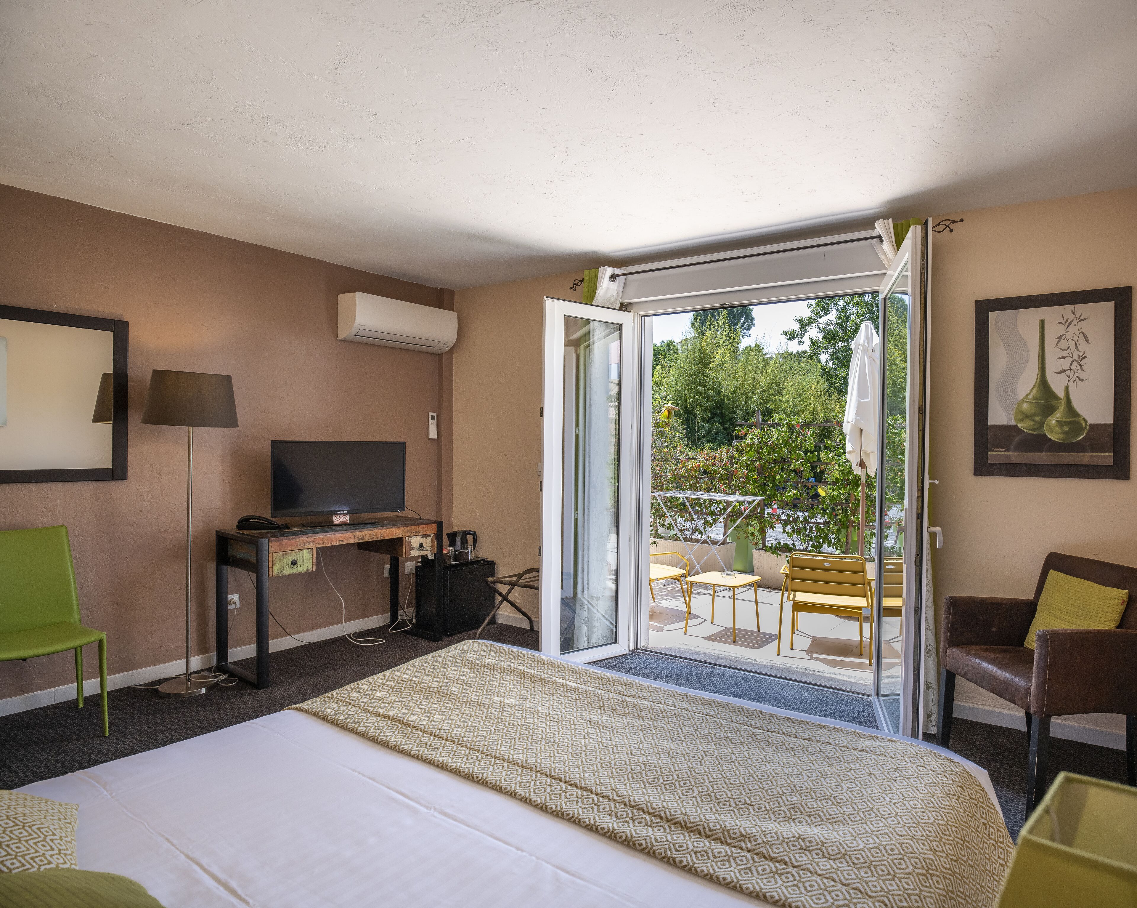 Habitación de hotel con cama, escritorio con televisor, silla y balcón con mobiliario exterior.