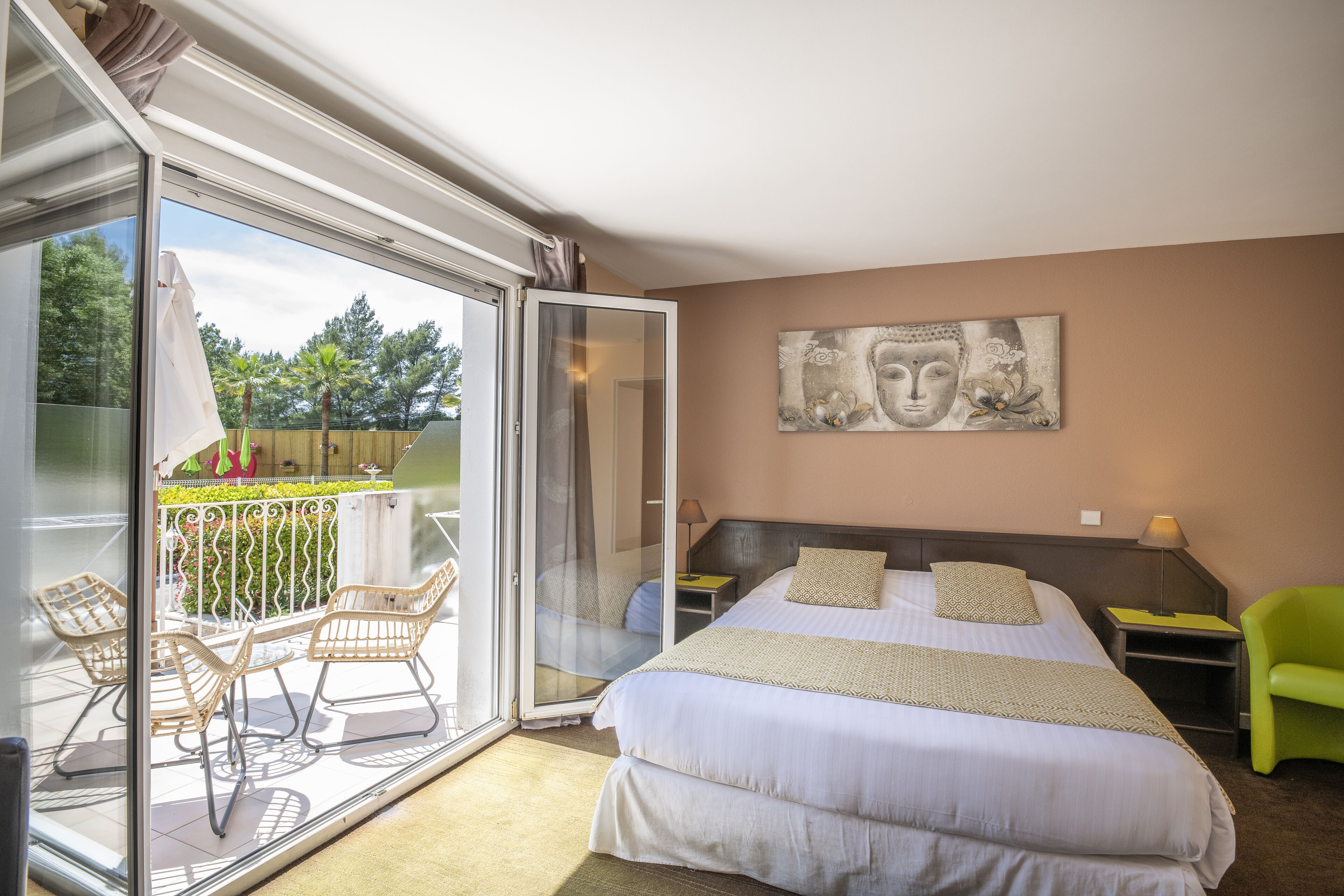 Ein Schlafzimmer mit einem großen Bett, einer Glastür, die auf einen Balkon mit Stühlen führt, und einem Buddha-Gemälde an der Wand.