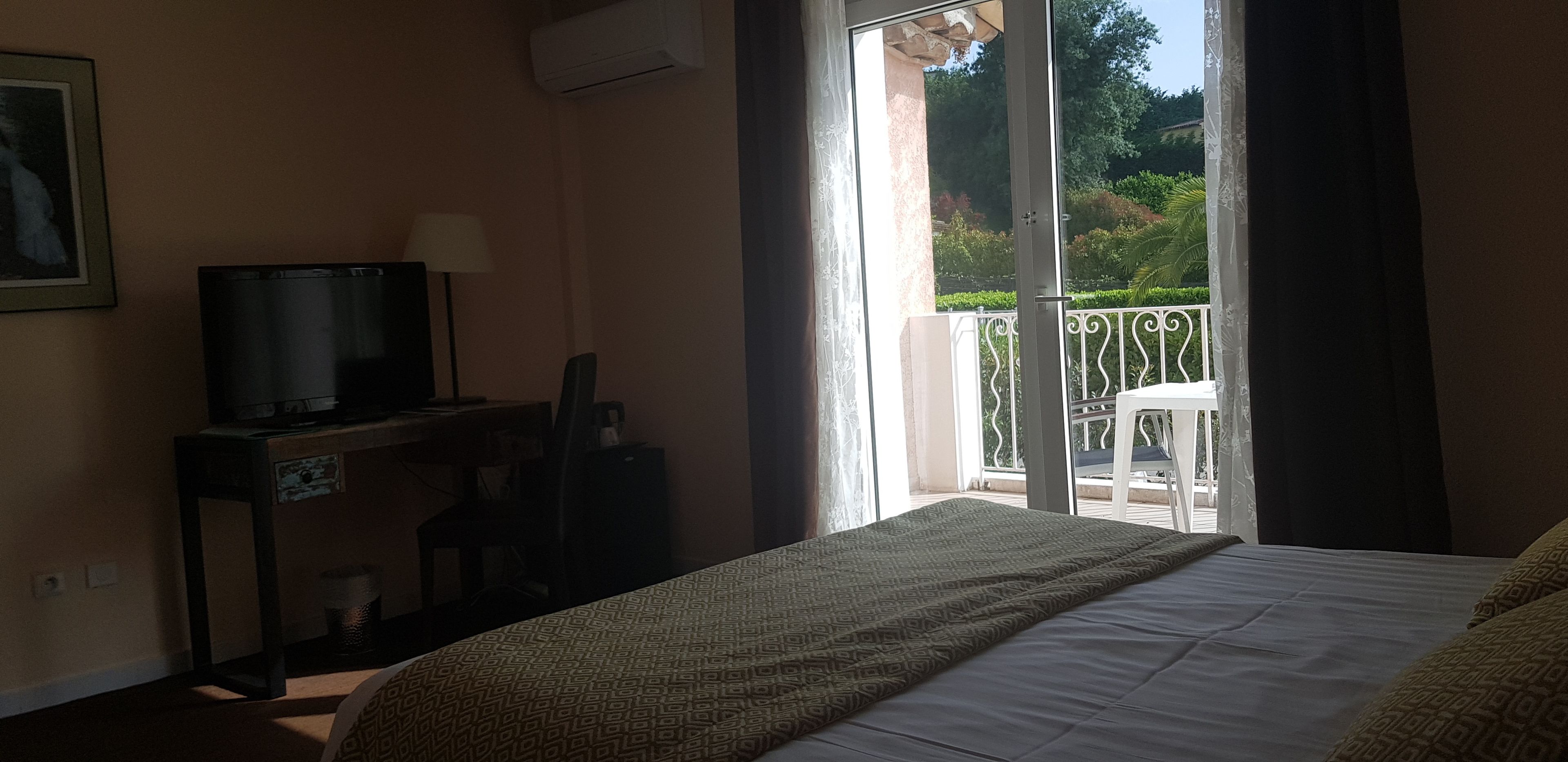 Une chambre d’hôtel comprenant un lit, un bureau avec une télévision, une chaise et un balcon donnant sur un espace verdoyant.