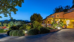 Un hotel bien iluminado con un camino de entrada pavimentado, rodeado de exuberante vegetación y con una montaña al fondo.