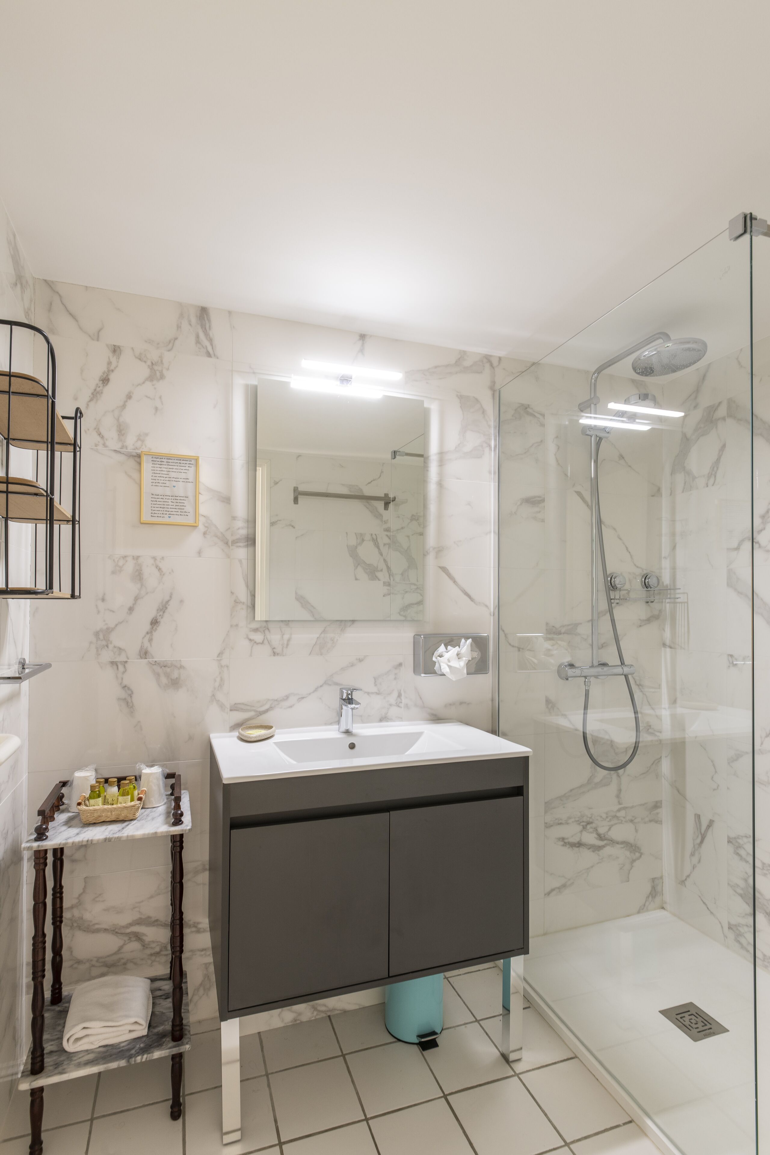 Un baño moderno con detalles de mármol en el salpicadero, un espejo grande, un lavamanos con toallero y una ducha con mampara de vidrio.