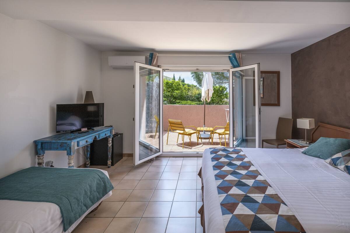 Ein Hotelzimmer mit Bett, Schreibtisch mit Fernseher, Balkon mit Stühlen und Blick auf Grünflächen.