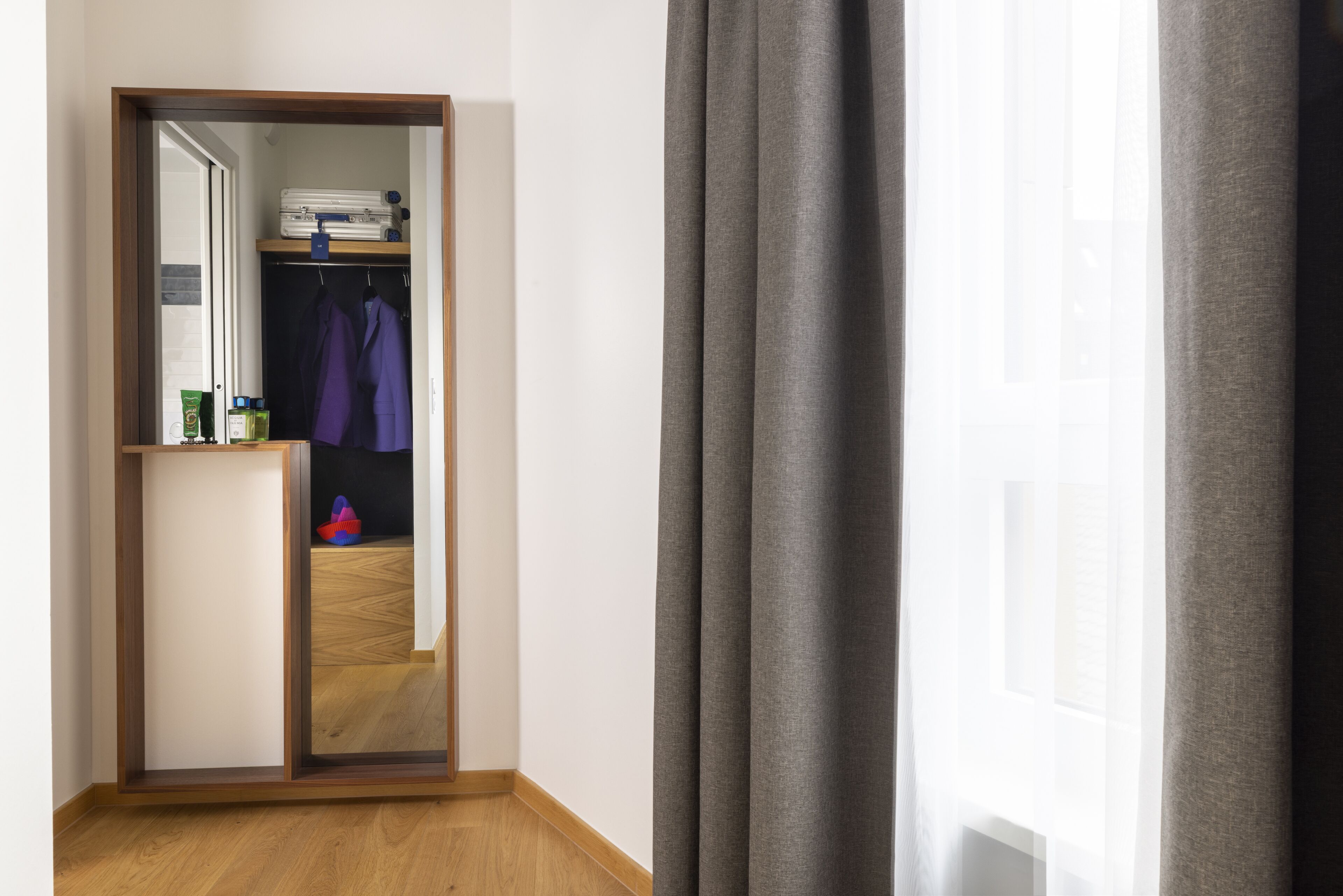 A hotel room with a wooden floor, a wardrobe with clothes, and a mirror reflecting the room.