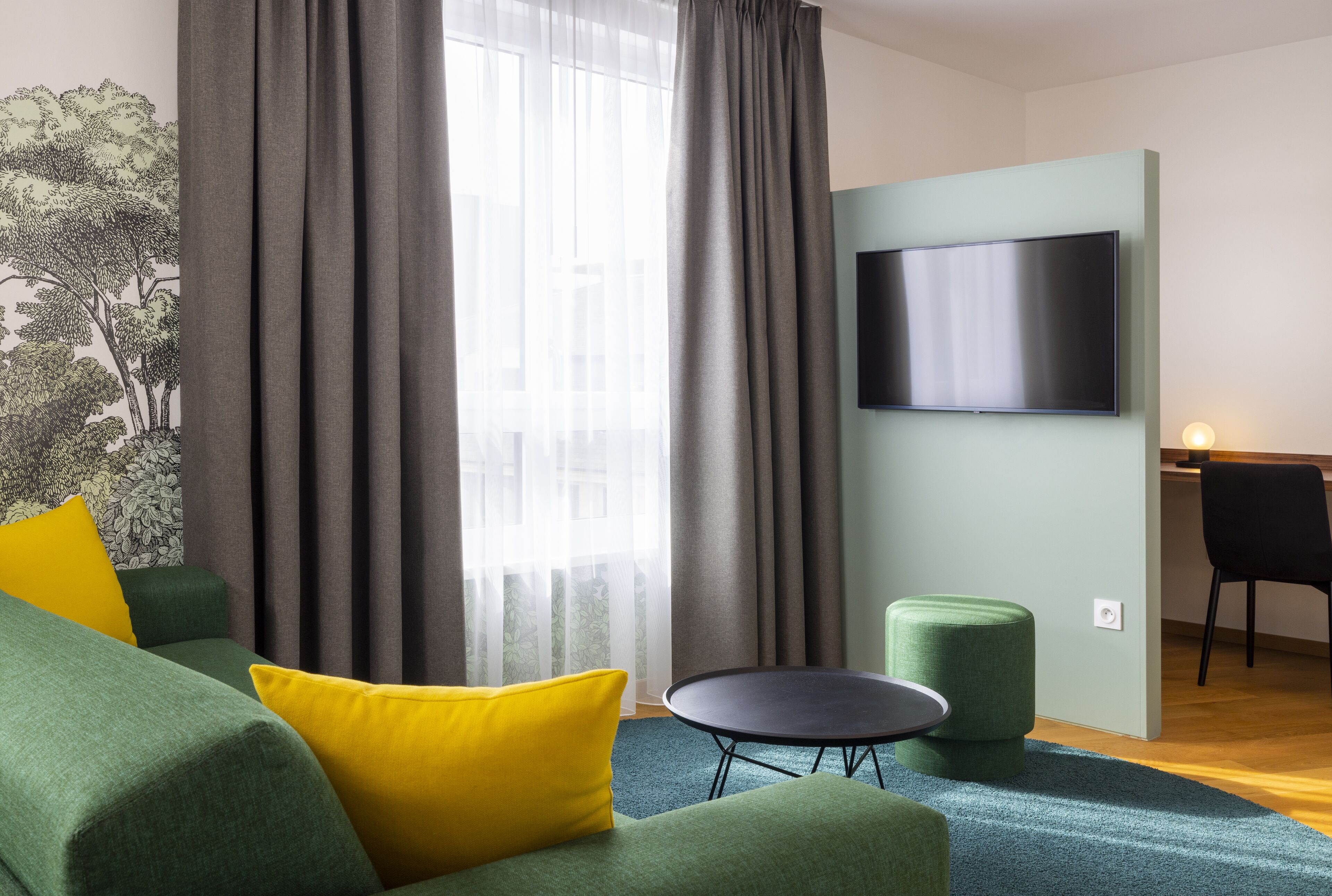 A modern living room with a green sofa, yellow pillows, a round coffee table, and a flat-screen TV mounted on the wall.