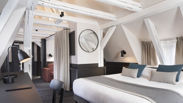 A modern hotel room with a large bed, a desk, and a chair. There is a round mirror on the wall and a window with curtains. - Hotel Du Dragon (Strasbourg)