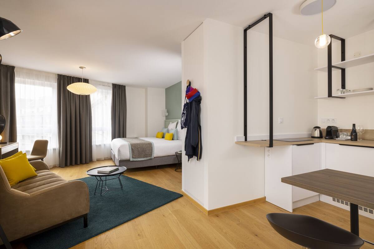 A modern hotel room with a bed, a sofa, a small dining table, and a kitchenette.