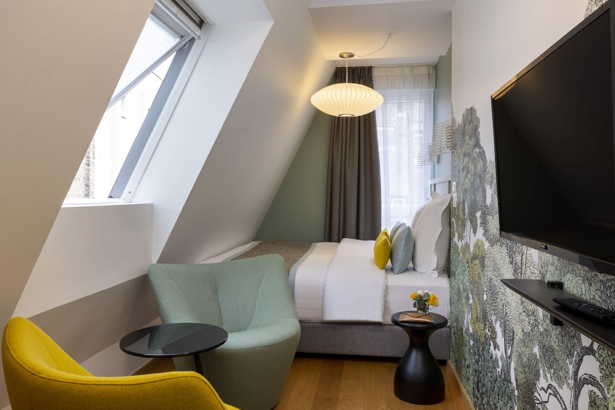 A modern hotel room with a bed, a television, a small table, a chair, and a window.