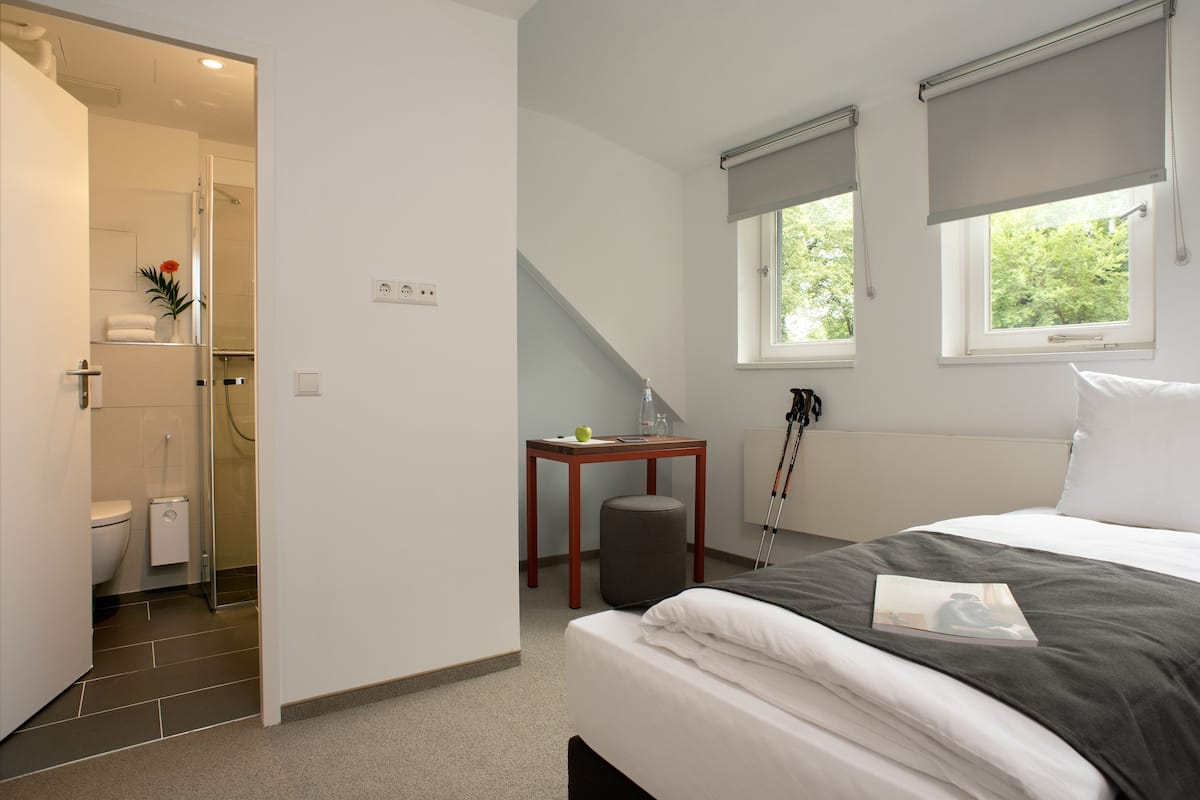 A bedroom with a bed, a bedside table, a window with a view, and a bathroom with a toilet and shower.