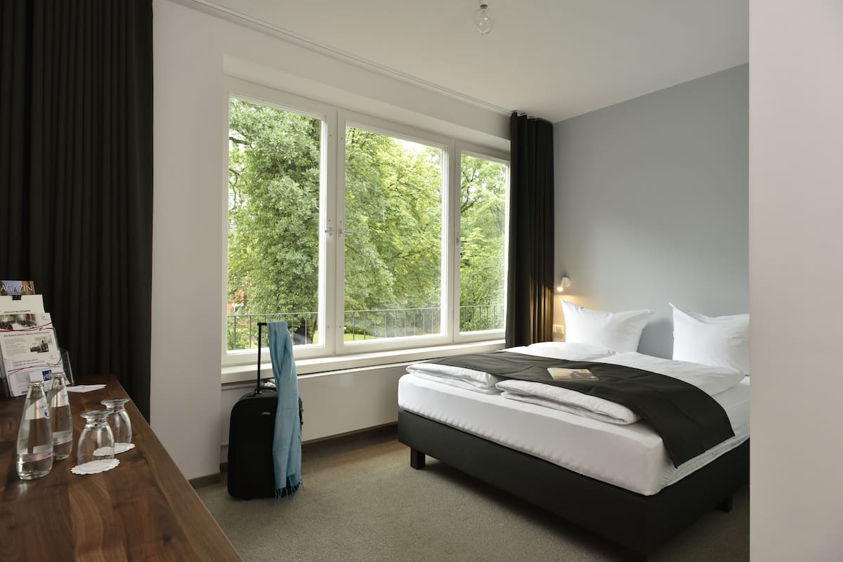 A hotel room with a large window, a bed, a desk with water bottles, and a suitcase.