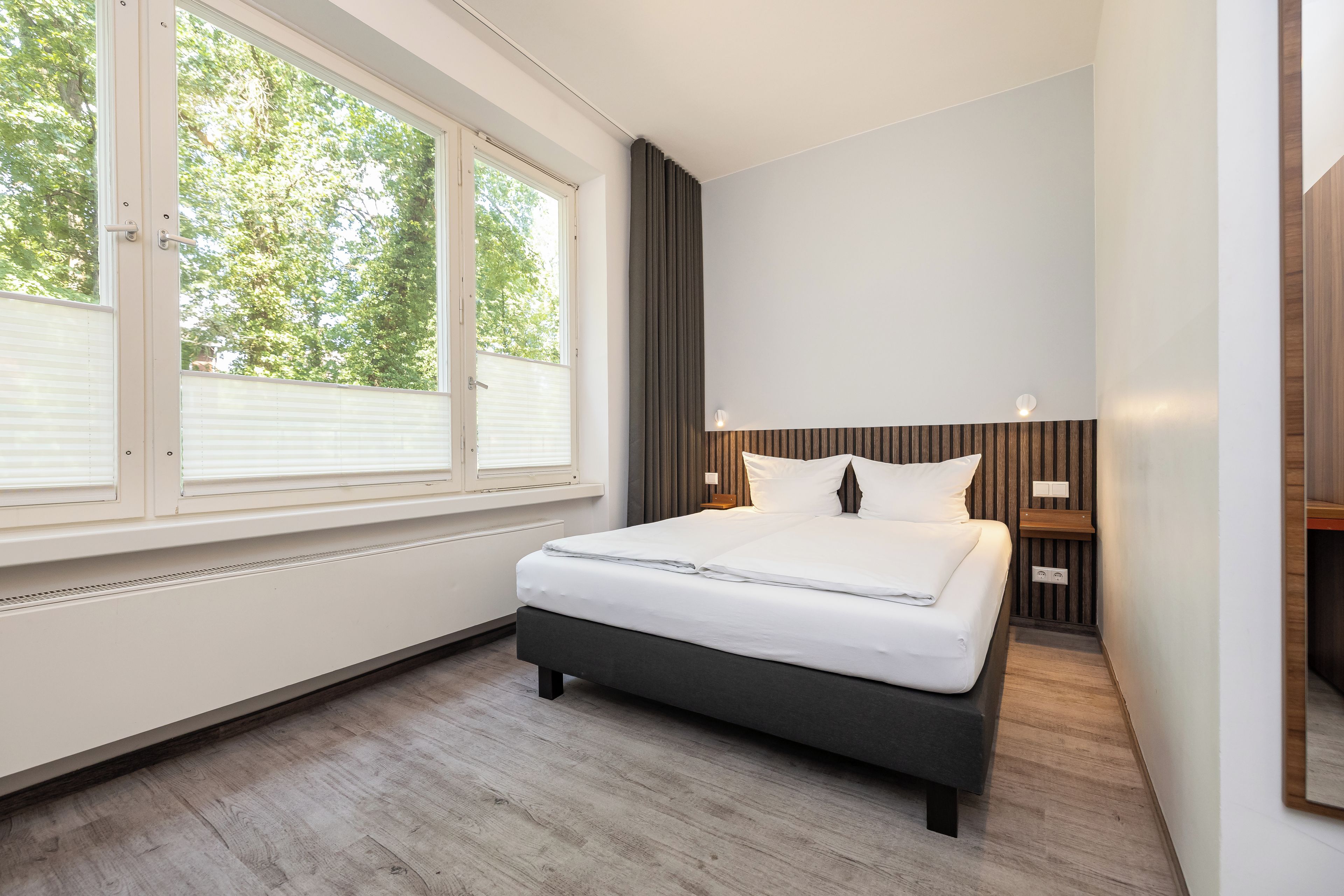 Un dormitorio moderno con un ventanal grande, una cama con sábanas blancas, un cabecero de madera y una mesita de noche con una lámpara.