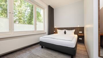 Un dormitorio moderno con un ventanal grande, una cama con sábanas blancas, un cabecero de madera y una mesita de noche con una lámpara.
