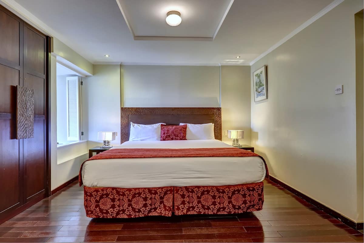 Une chambre d’hôtel avec un grand lit, une tête de lit en bois, une table de chevet, une lampe et une fenêtre avec des stores.