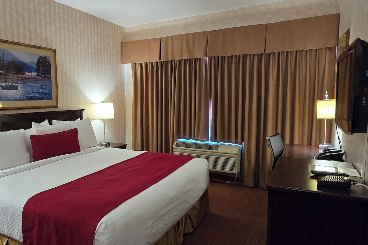 Une chambre d’hôtel avec un grand lit, un tableau au mur, un bureau avec un ordinateur et une fenêtre avec des rideaux.