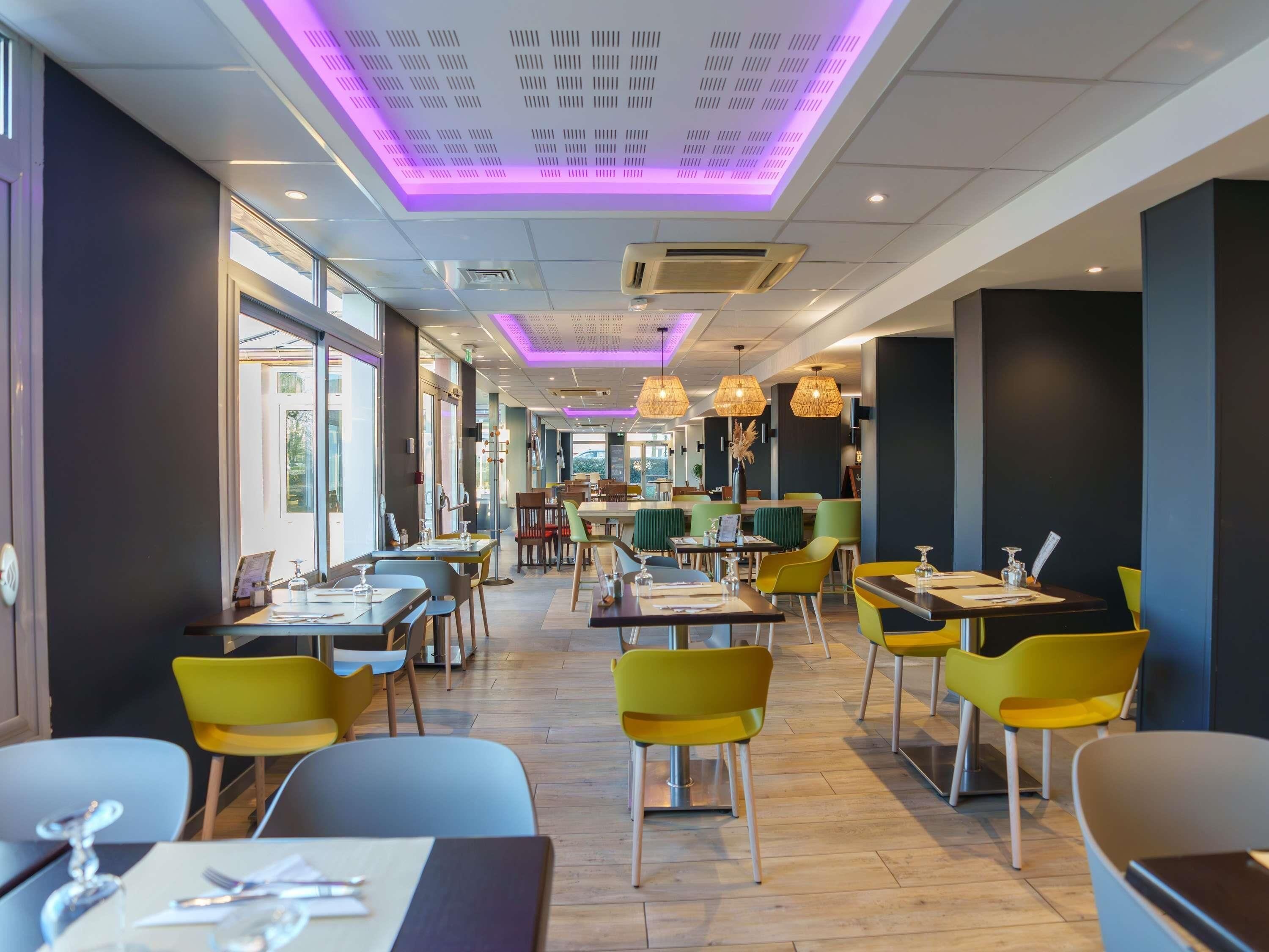 A modern restaurant interior with wooden flooring, yellow and green chairs, and purple ceiling lighting.