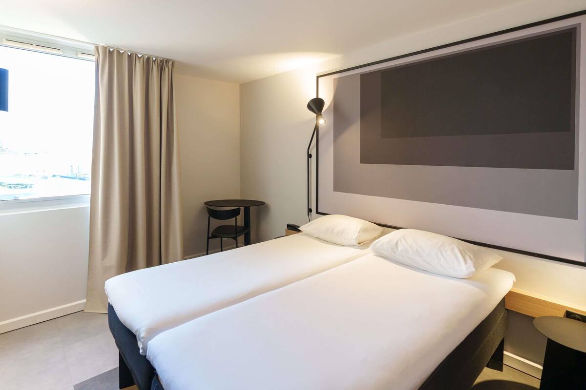 A hotel room with a large bed, a small table, a chair, and a large window with curtains.
