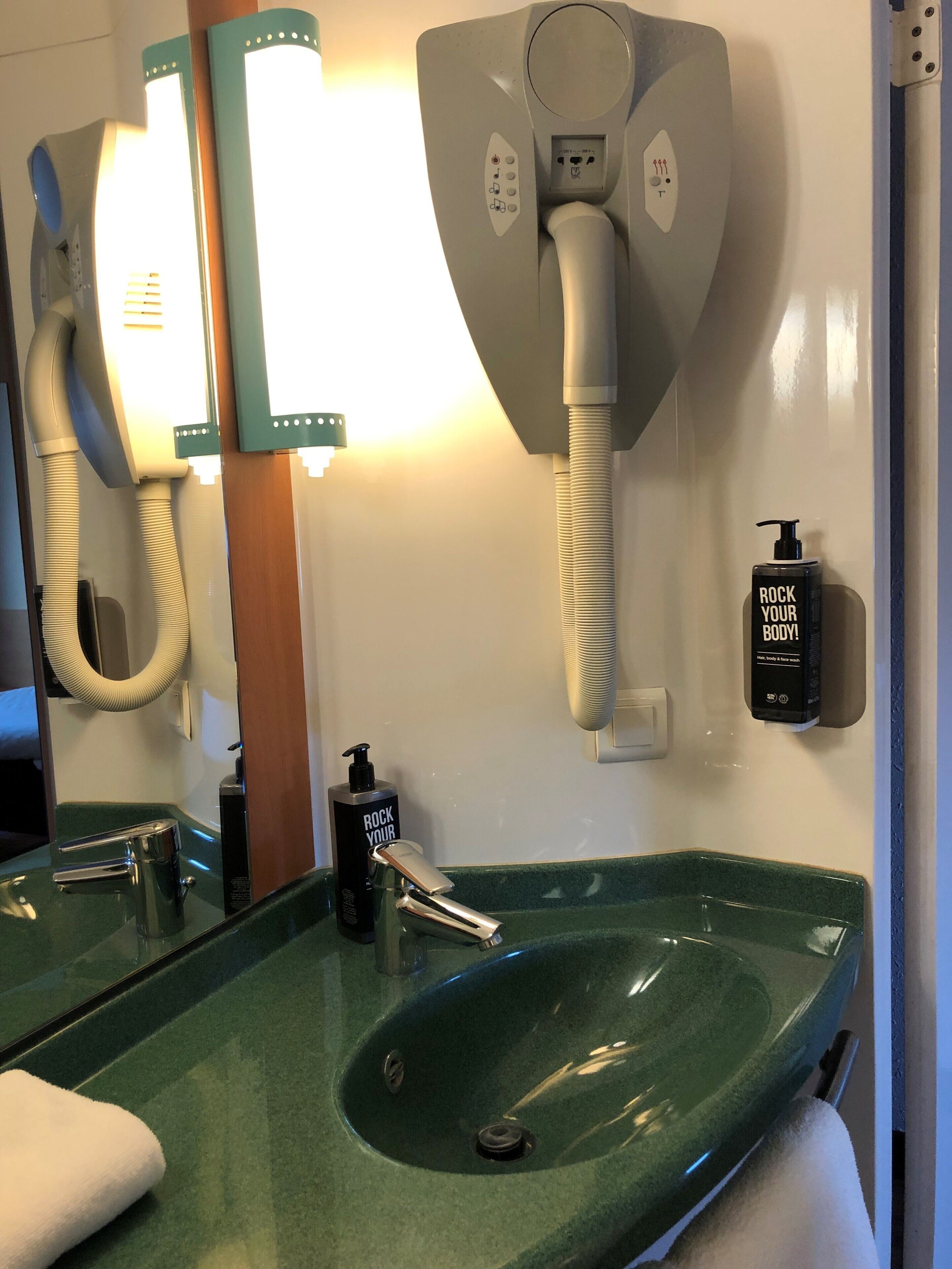 Un baño con un lavamanos verde, dos espejos, un secador de pelo y una botella de loción corporal.