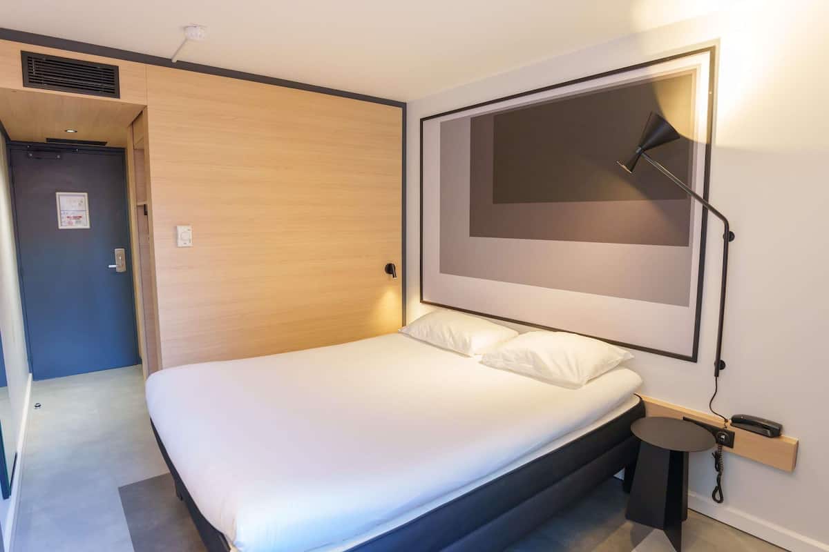 A modern bedroom with a bed, a bedside table with a lamp, and a door leading to another room.