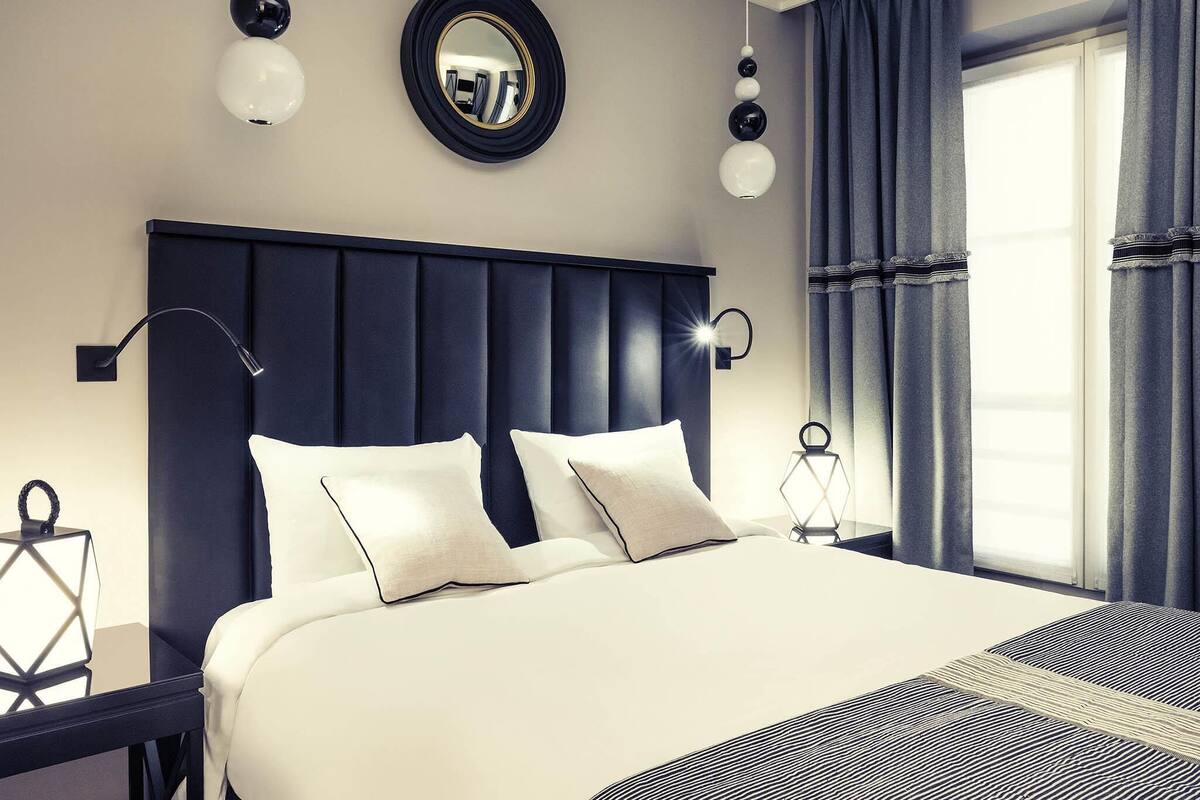 Una habitación de hotel moderna con una cama grande, cabecero oscuro y un ventanal con cortinas.