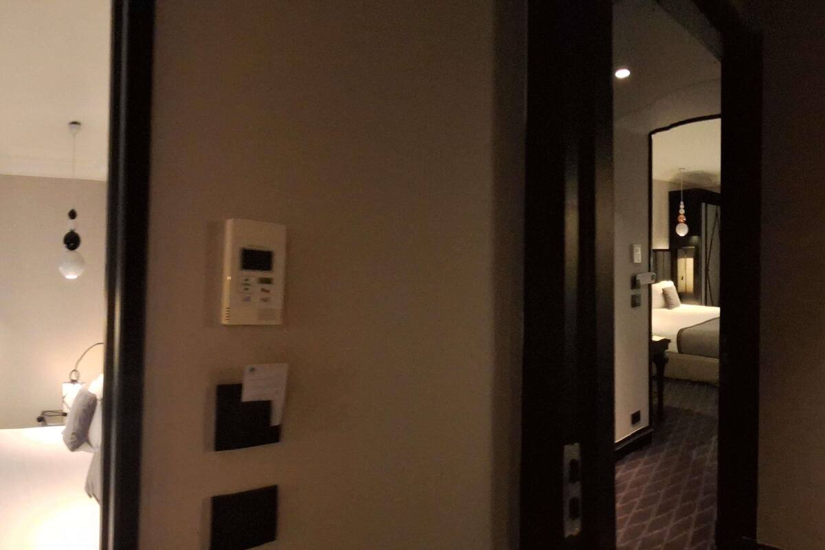 Habitación de hotel con cama, mesita de noche, espejo y termostato en la pared.