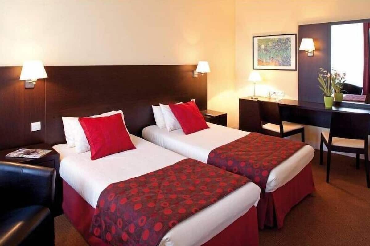 Une chambre d’hôtel avec deux lits, chacun agrémenté de draps rouges et blancs, un bureau avec une chaise et une grande fenêtre avec des rideaux.