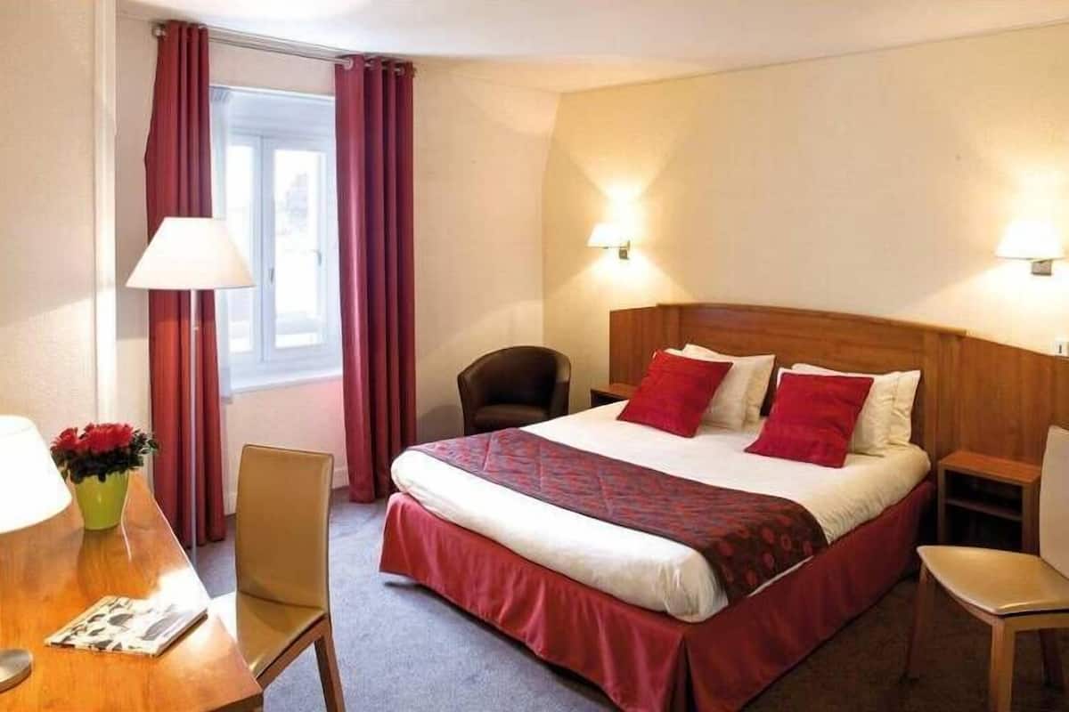 Une chambre d’hôtel avec un lit, un bureau avec une lampe, une chaise et une fenêtre avec des rideaux.