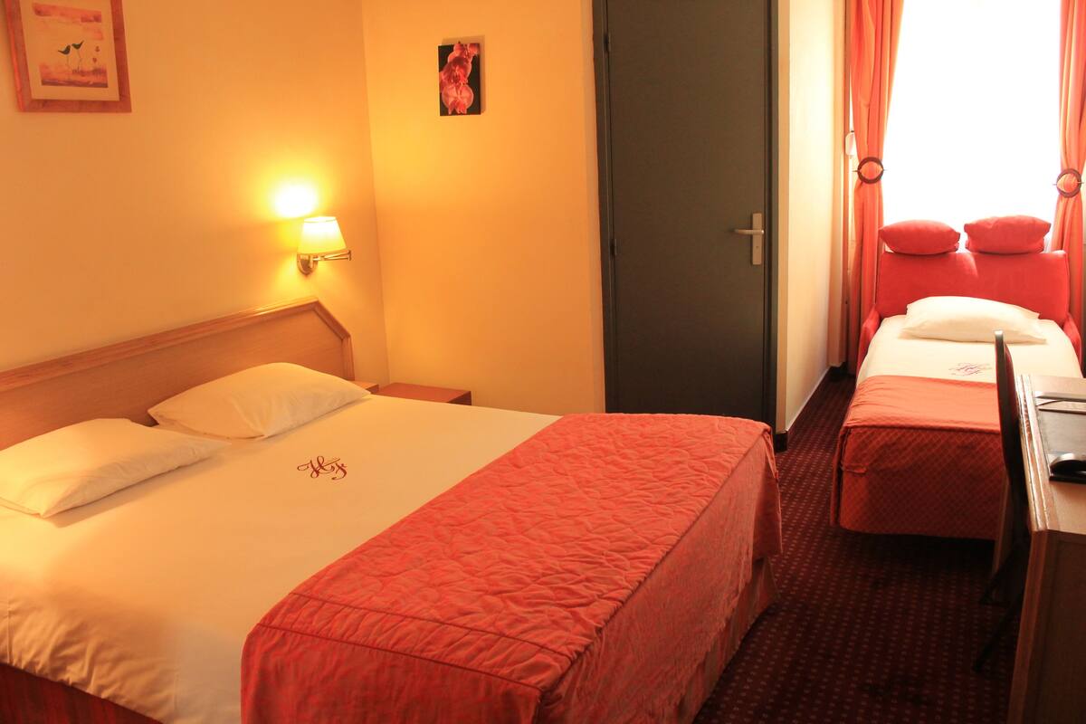 A hotel room with a large bed, a smaller bed, a desk, and a chair. There is a door and a window with curtains.