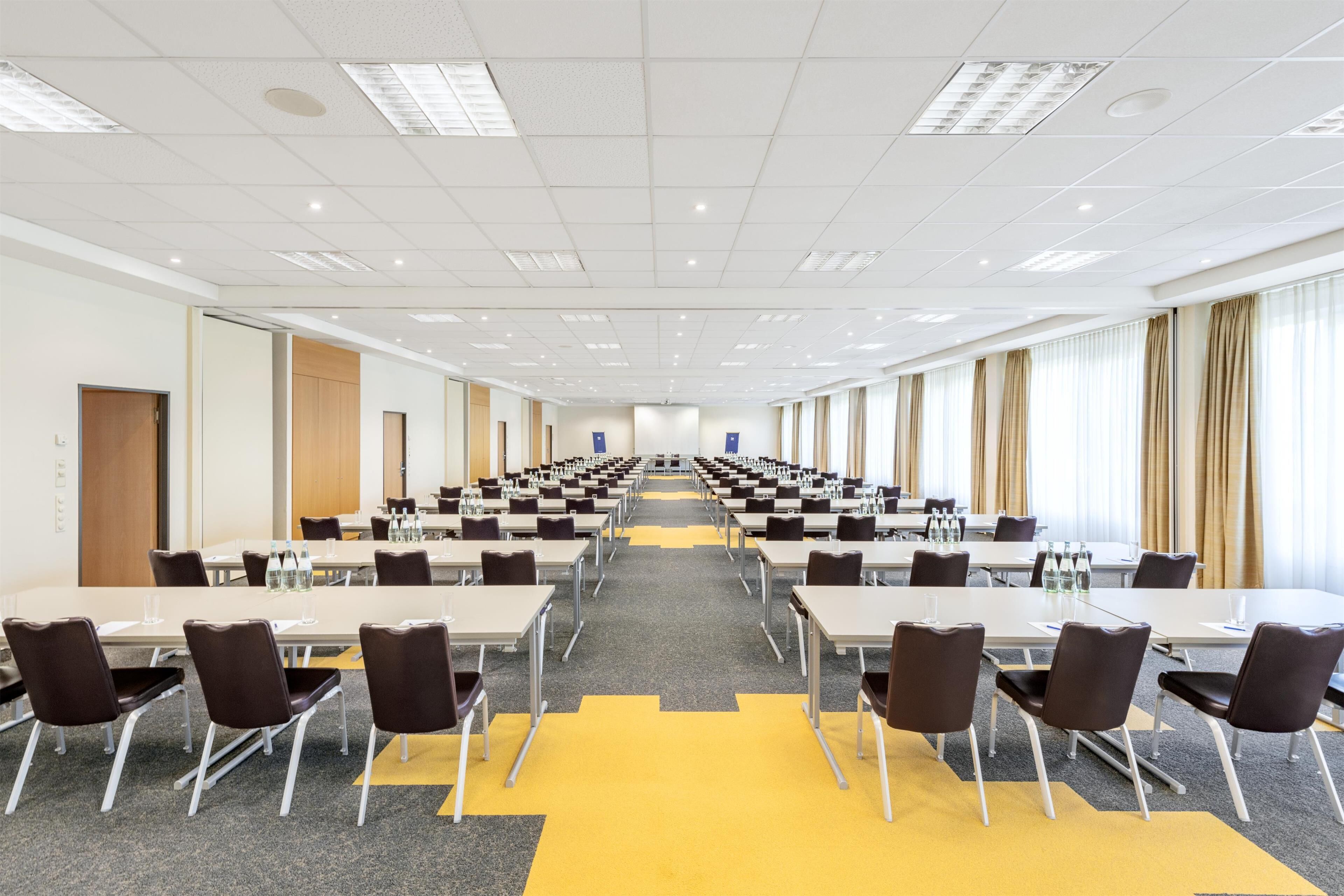 A conference room with multiple tables and chairs, arranged in rows, with a carpeted floor and a ceiling with recessed lighting.