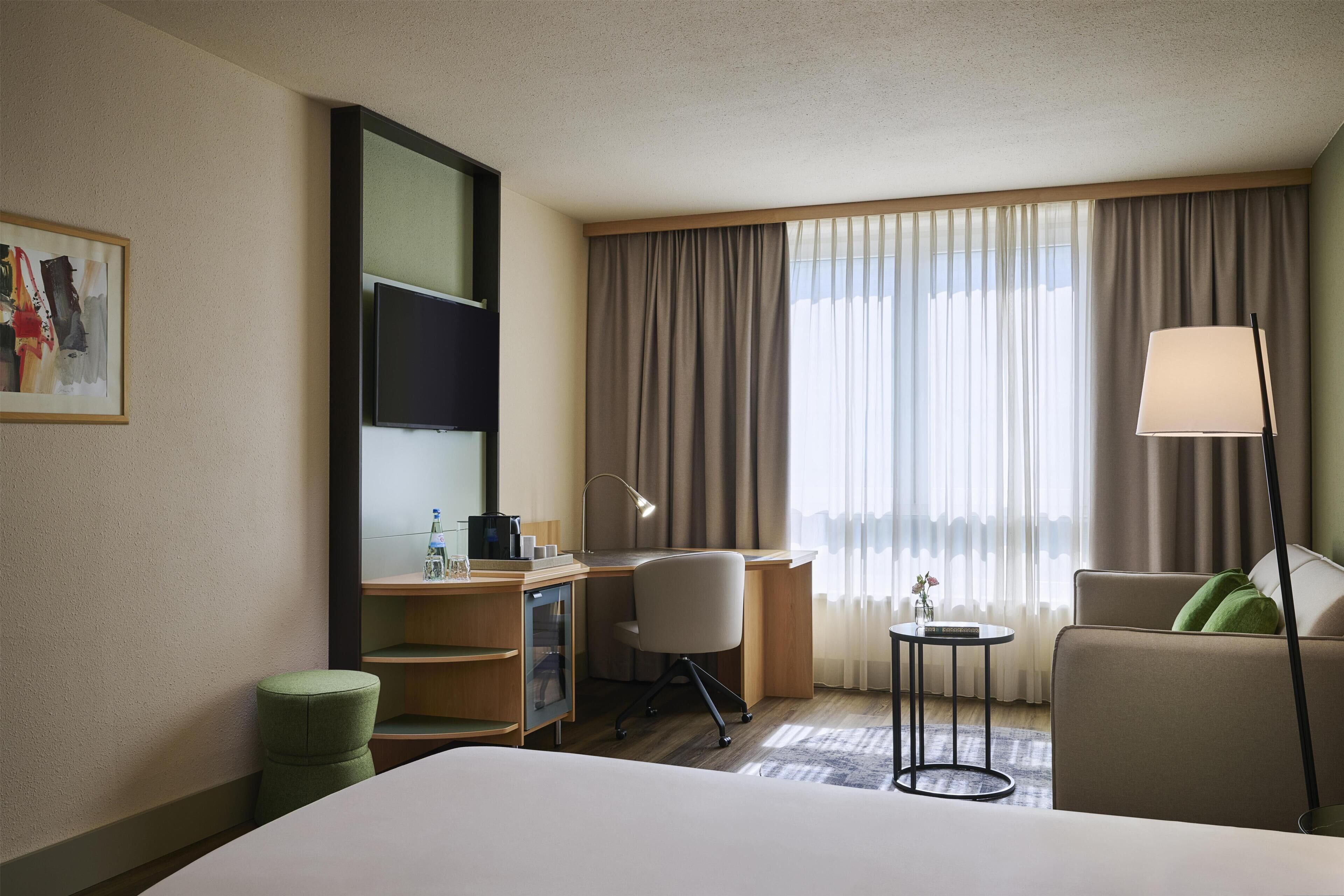 A hotel room with a bed, desk, chair, lamp, and a view of the outdoors through the window.