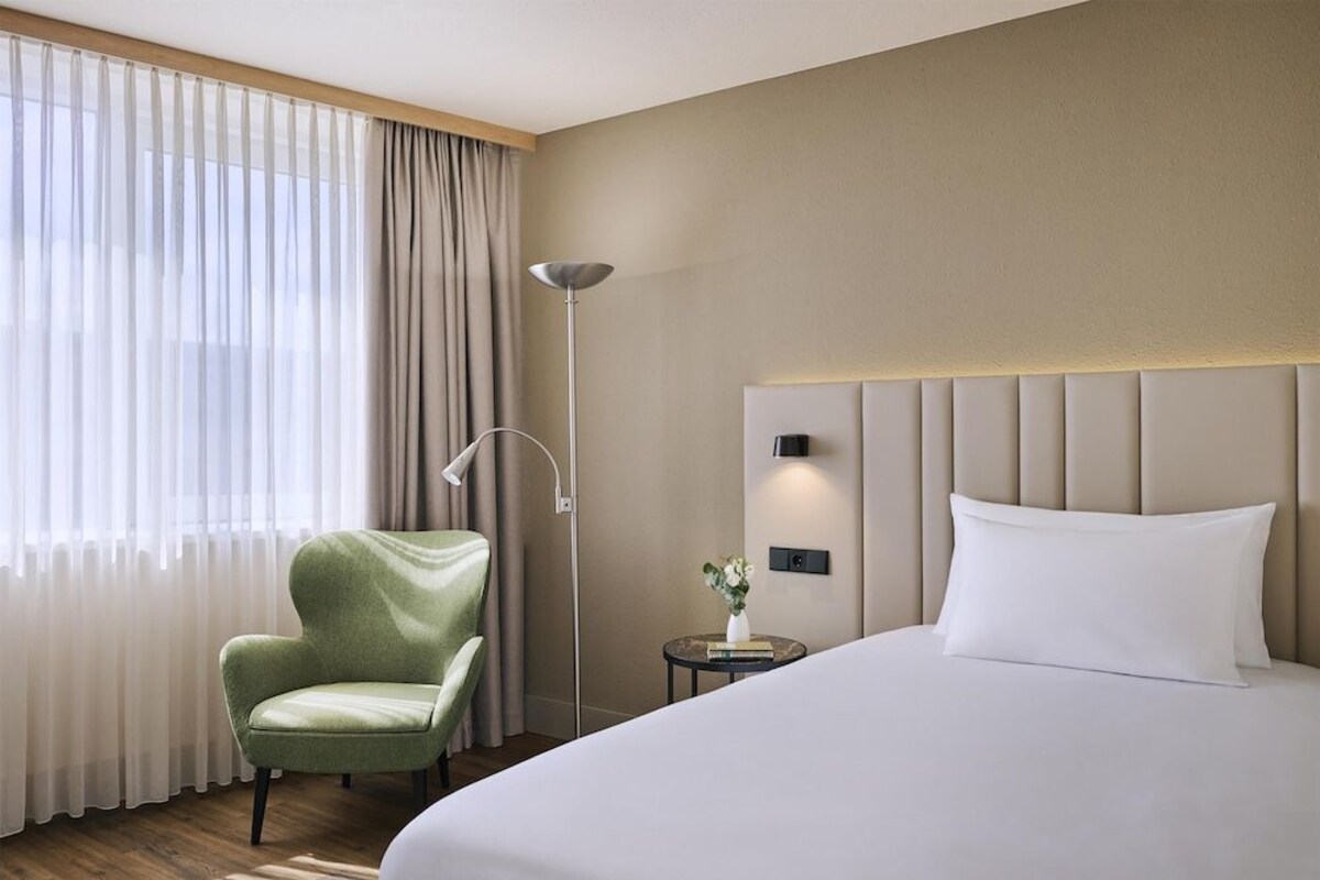 Habitación de hotel con una cama, un sillón verde, una lámpara de pie, una mesita con un florero y vistas a través de cortinas de gasa.